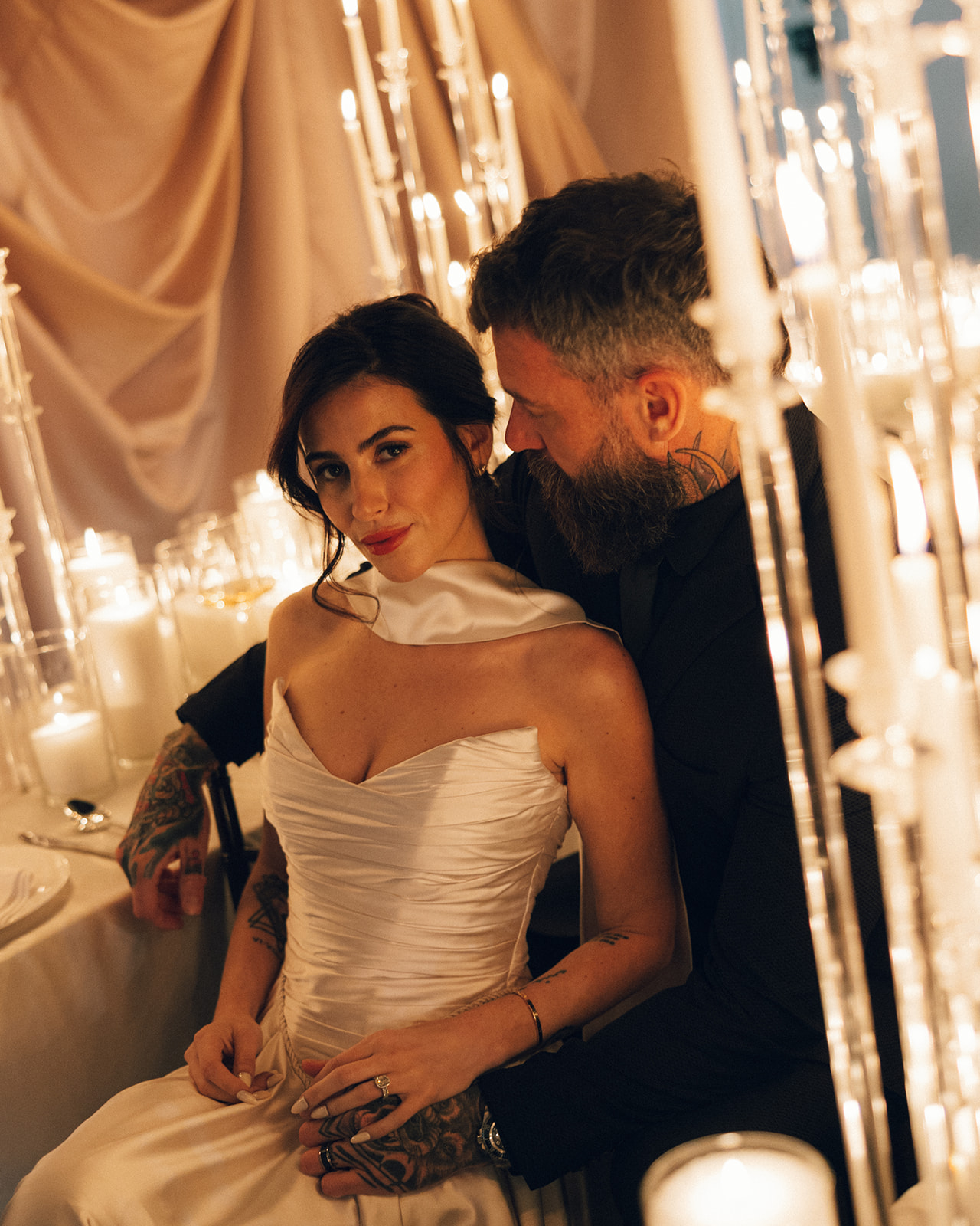 A woman in a white dress and a man with a beard sit close together in a dimly lit setting with candles and drapes.
