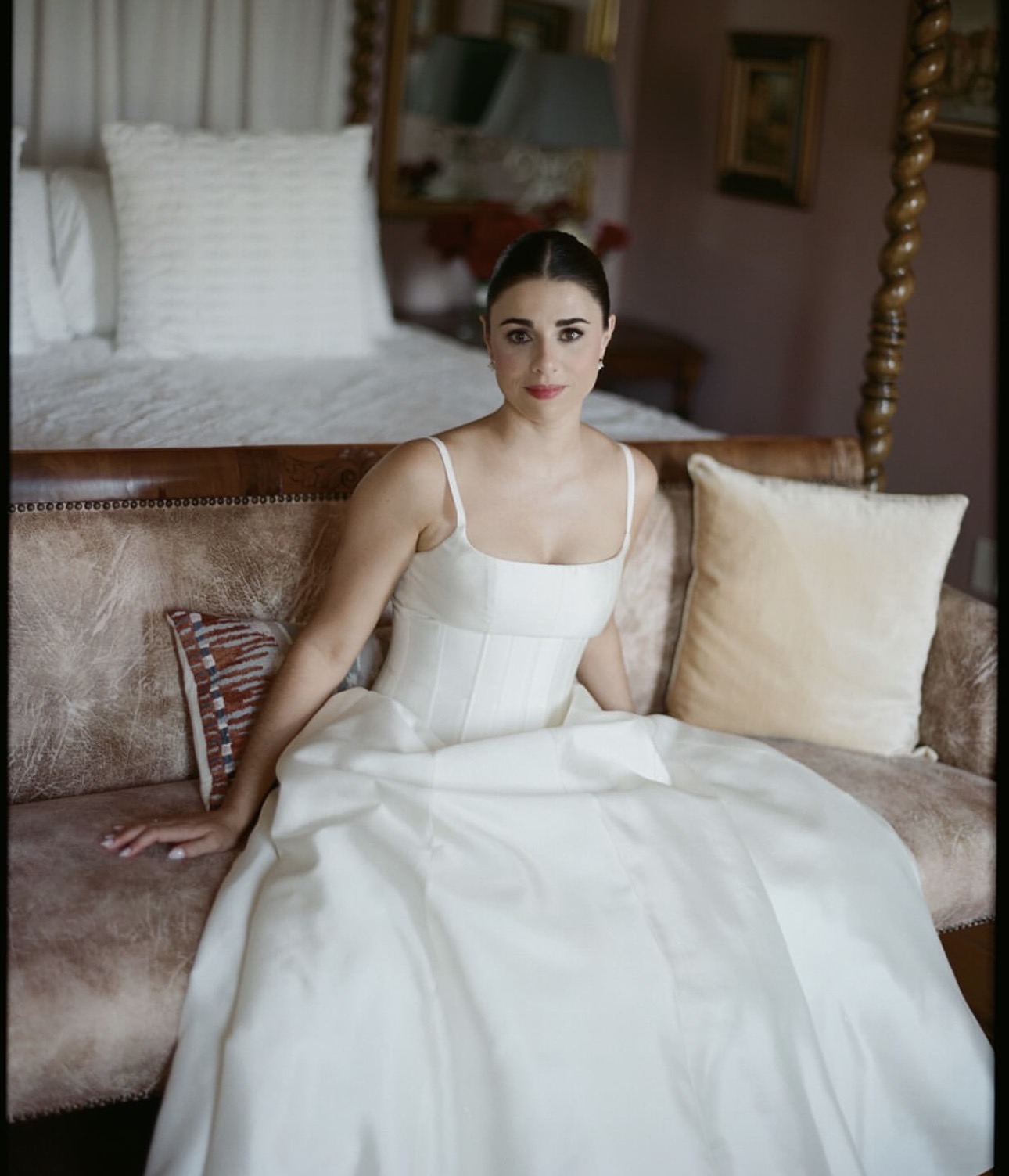 Woman in white dress sitting on a sofa in a room with bed and pillows behind her.
