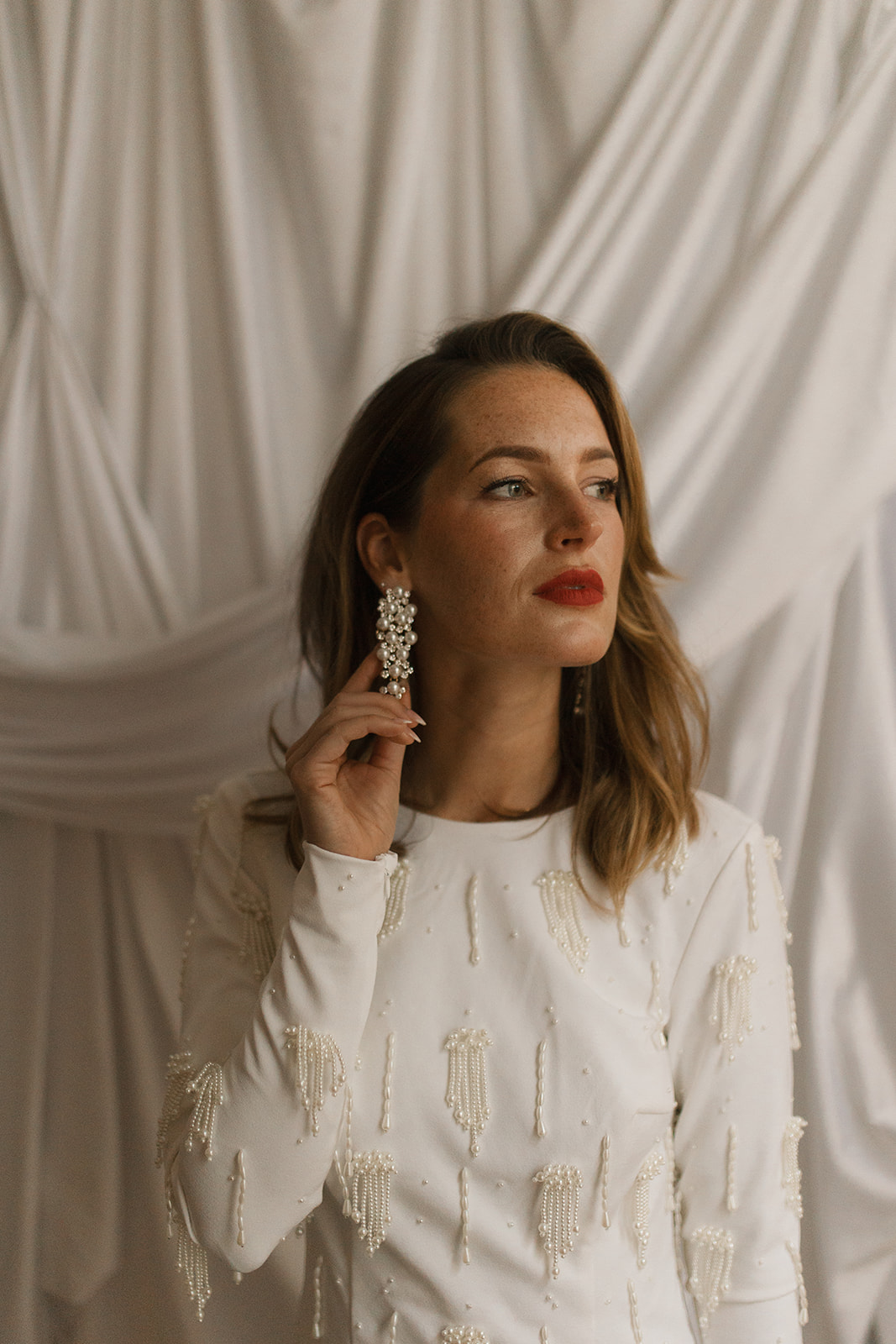 Woman with brown hair and red lipstick wearing large earrings and a white textured top, standing in front of draped fabric.