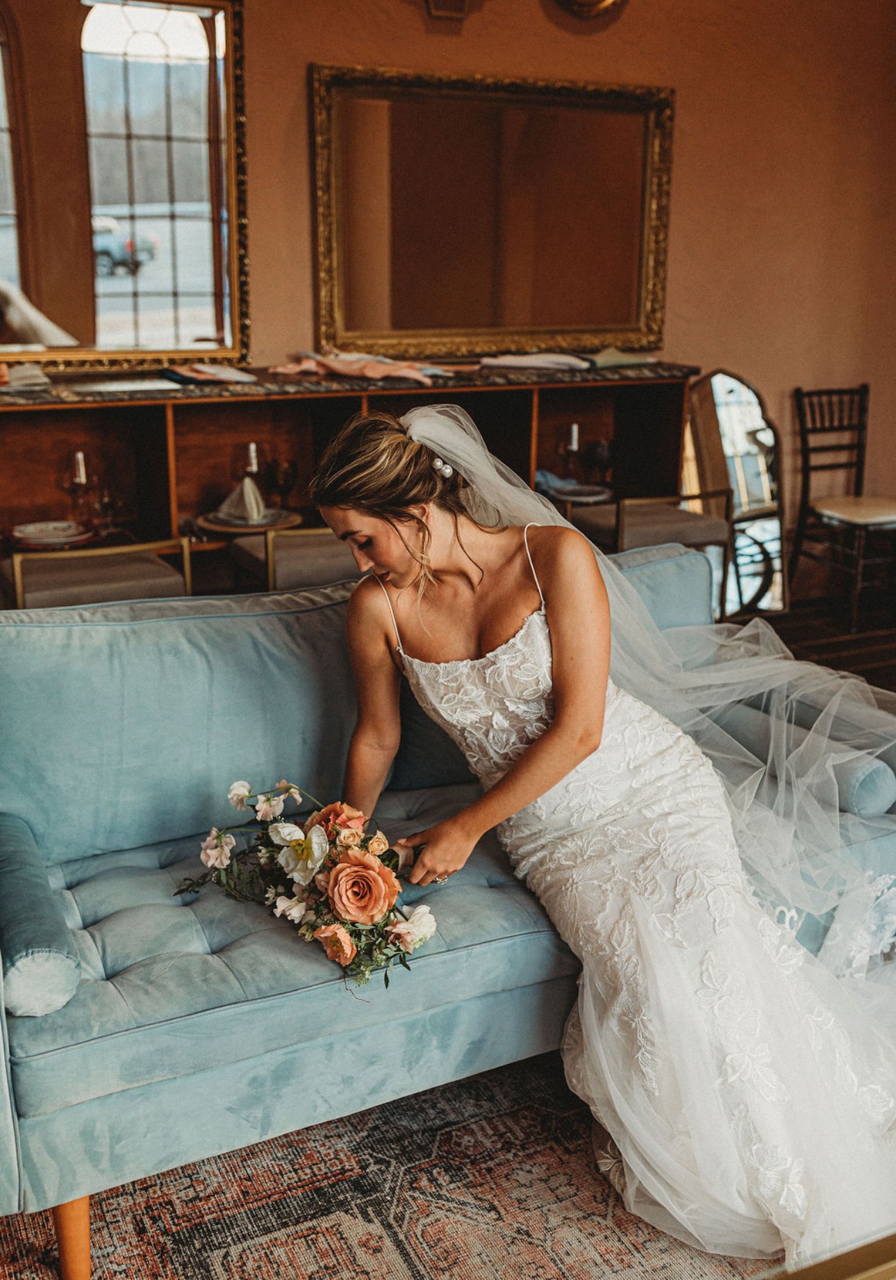 Bride in wedding dress arranging a bouquet on a blue sofa in a room with mirrors and wooden furniture.