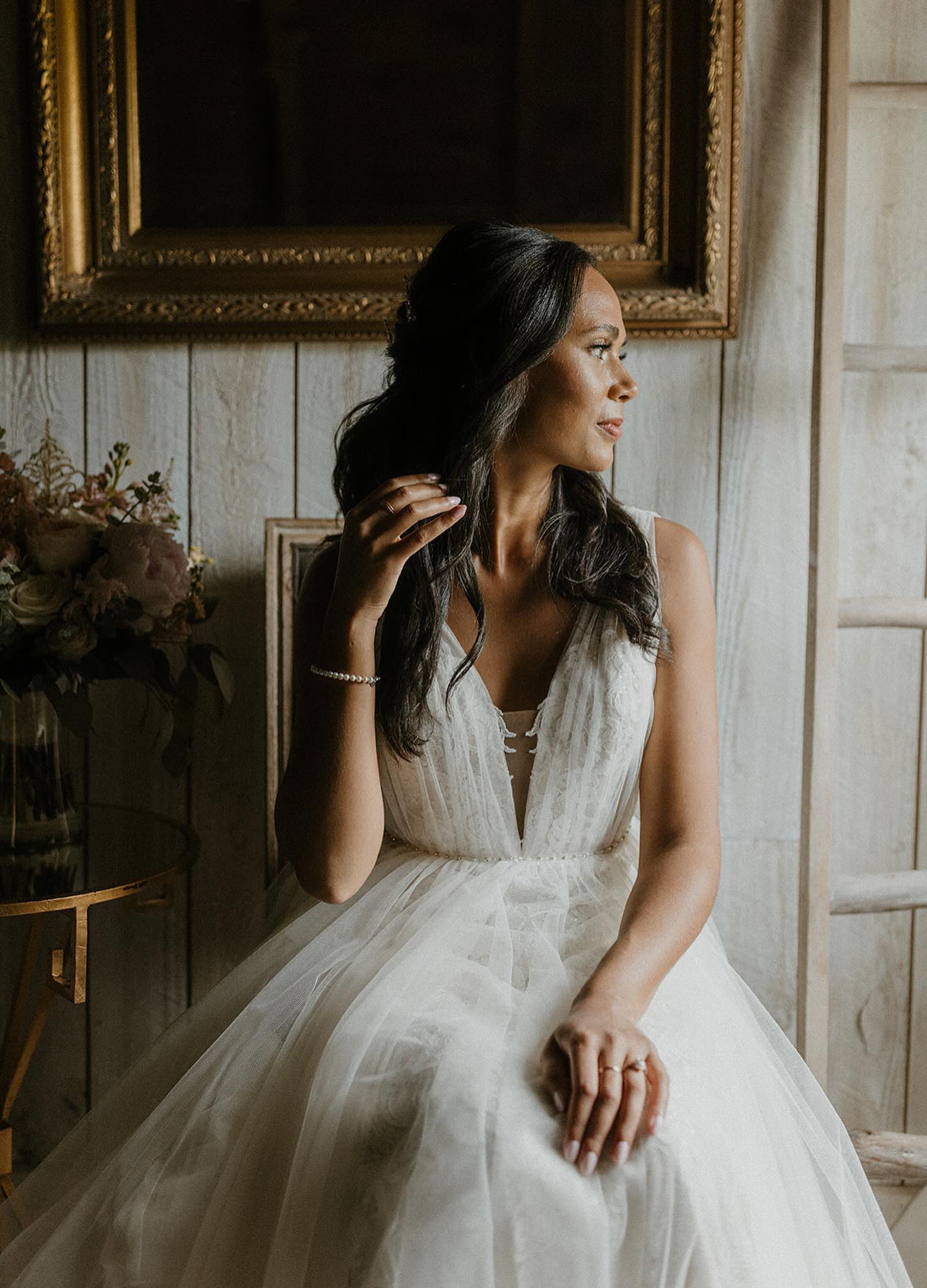 Woman in a white dress sitting by a window, looking to the side, with a large framed painting behind her.