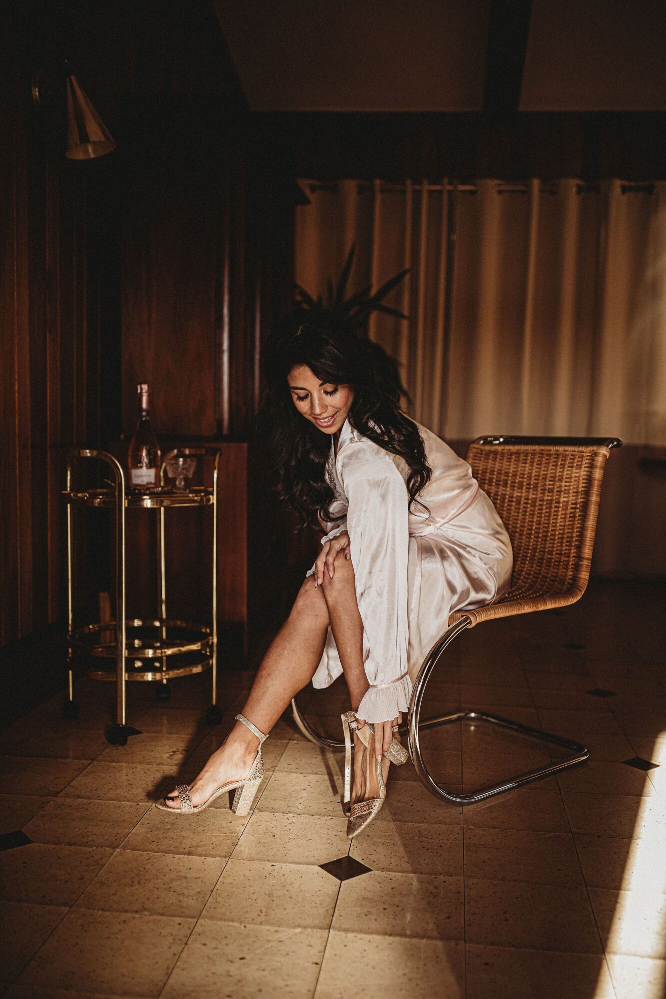 Woman in white dress sitting on a chair, adjusting high-heeled shoes, in a dimly lit room with curtains and wooden walls.
