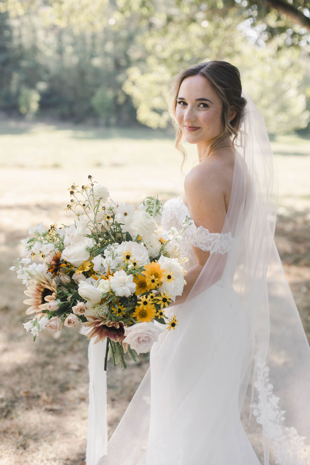 Bride in wedding dress holding a large bouquet outdoors, smiling, with trees and sunlight in background.