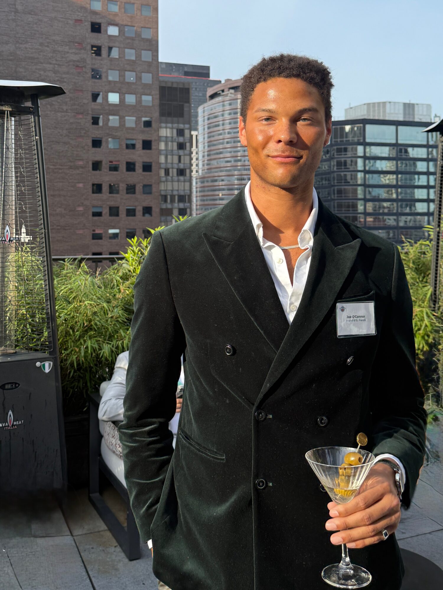 Young man in a black suit holding a cocktail, standing outdoors with city buildings in background.