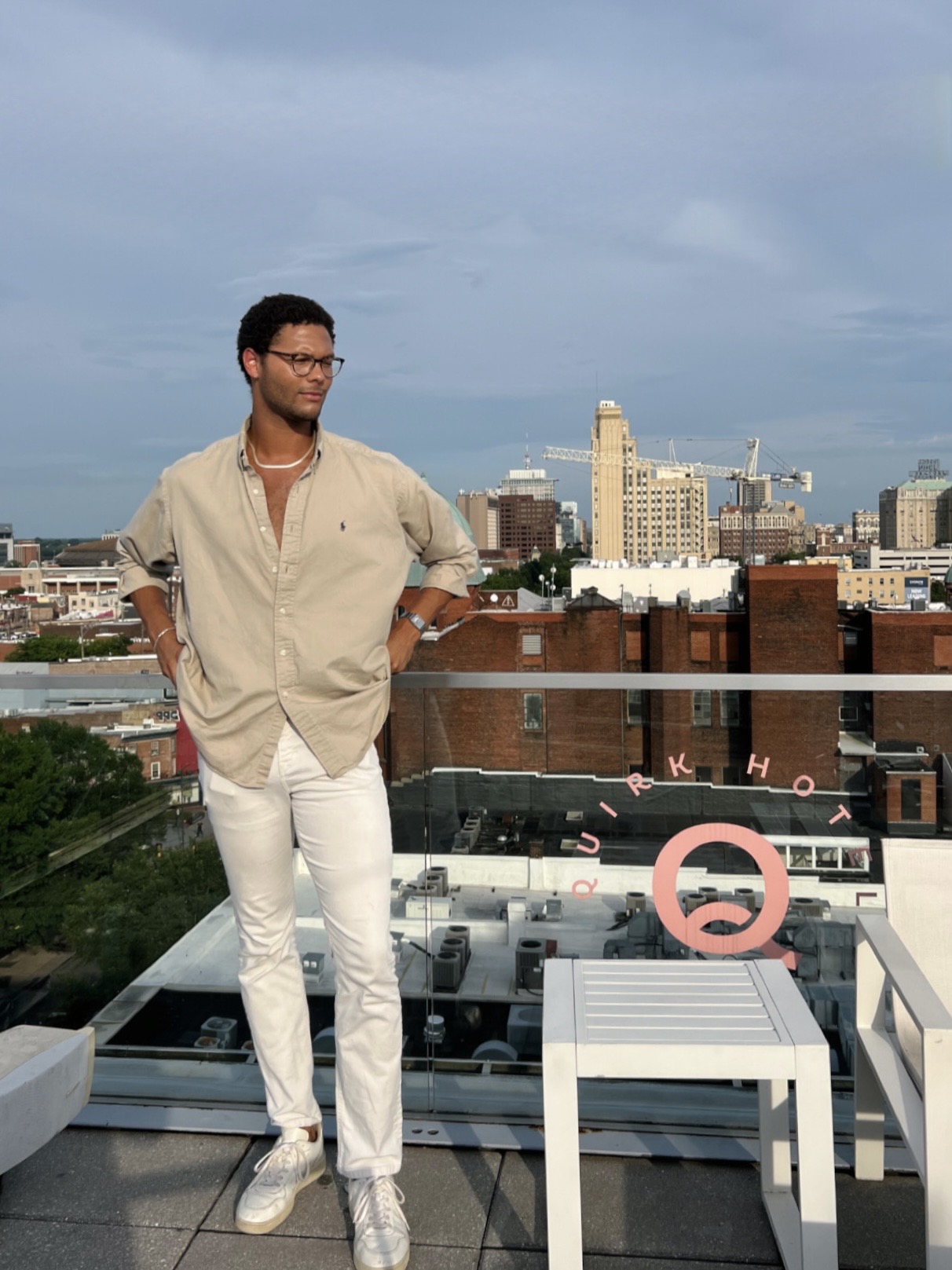 Man with glasses and beige shirt standing on rooftop with city skyline in background, white table nearby, cloudy sky.