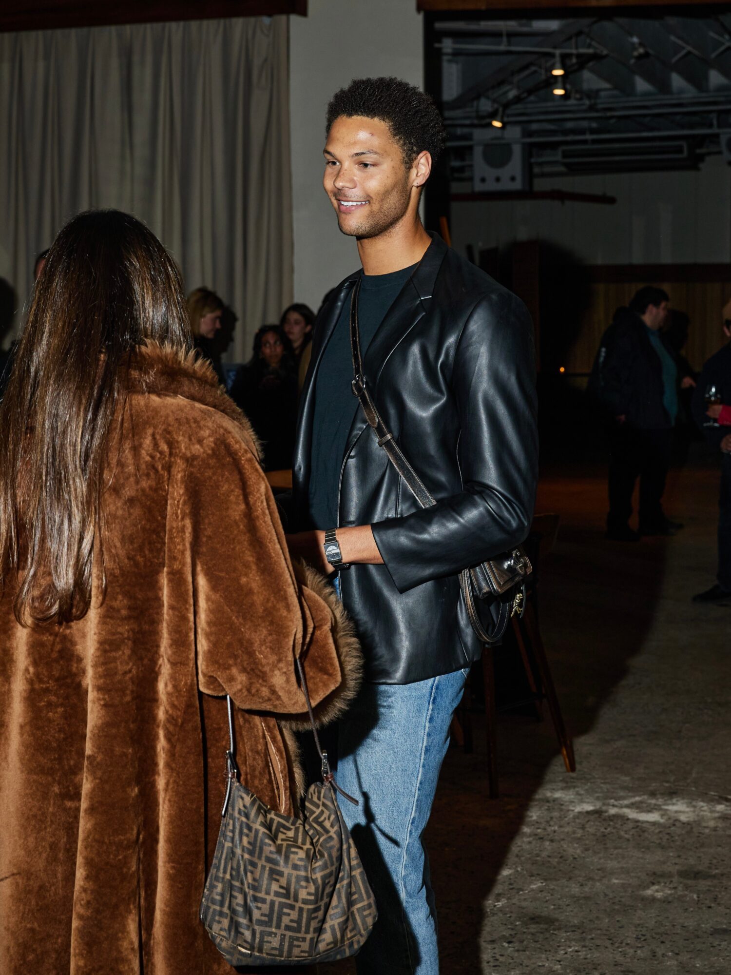 Young man smiling and talking to a woman in a brown coat at an indoor event.
