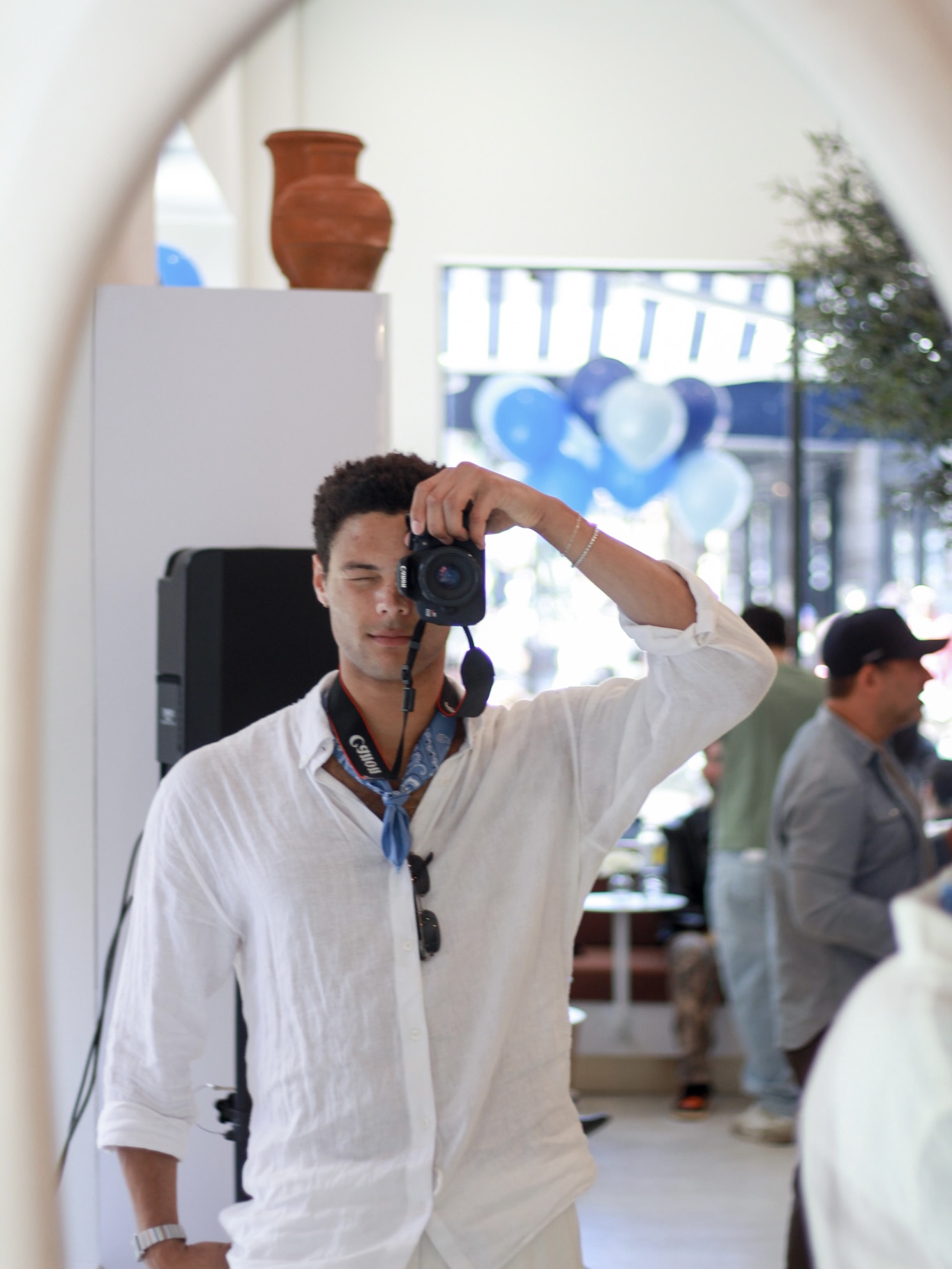 Young man taking a selfie in a mirror, wearing a white shirt, with a camera in hand, indoors with other people and decor visible.