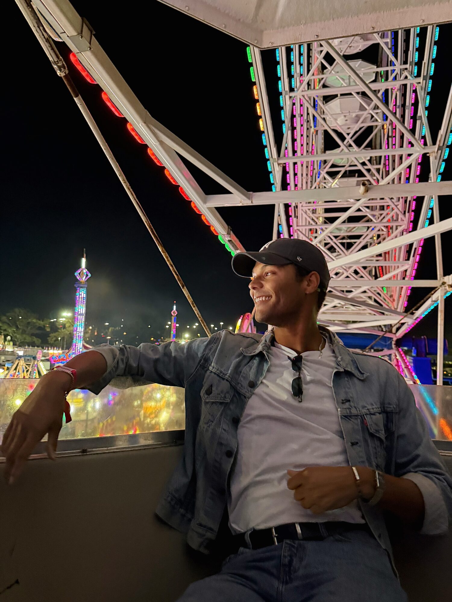 Man wearing a cap and denim jacket sitting on a ferris wheel ride at night, illuminated amusement park in background.