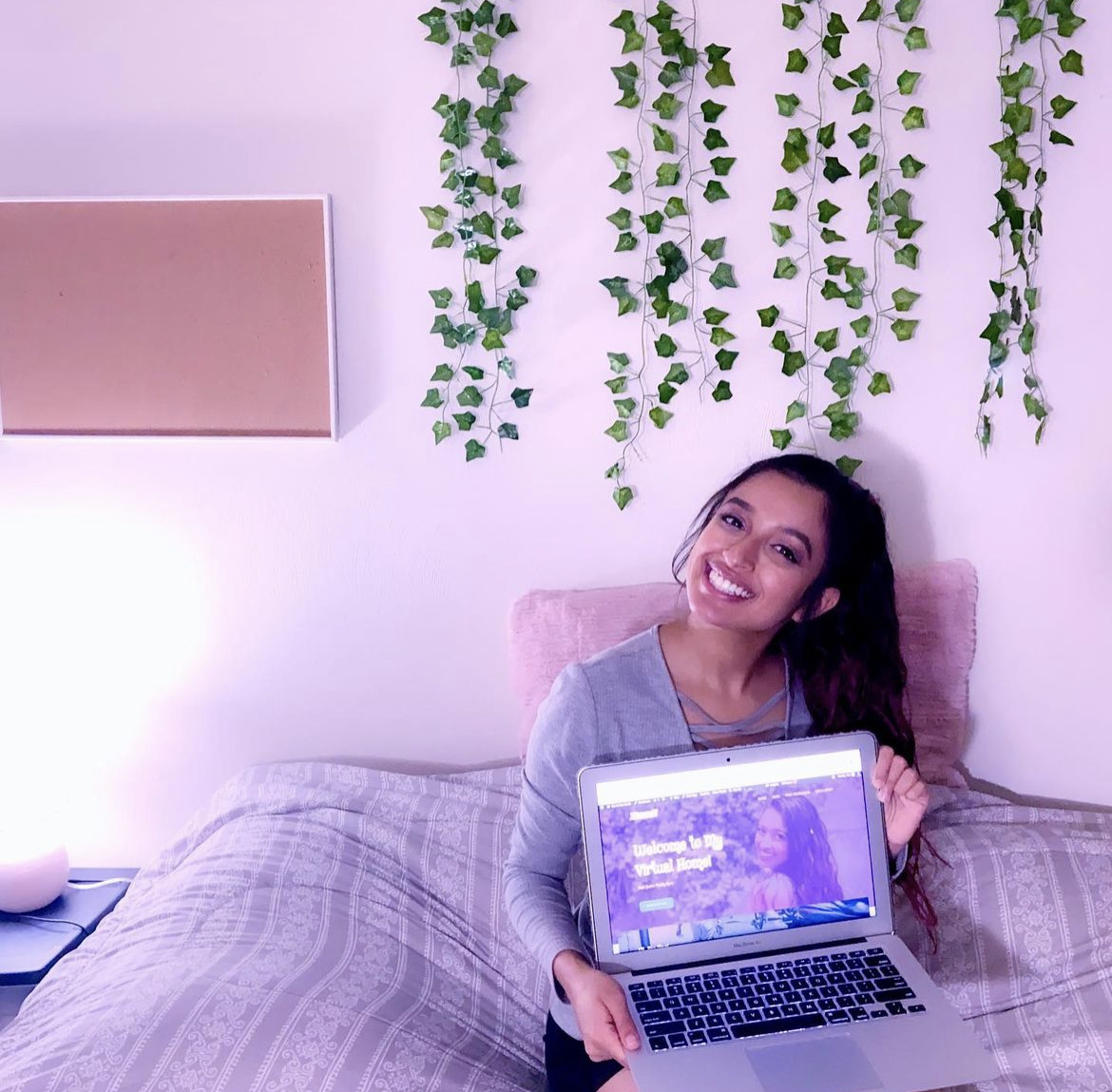 Woman sitting on bed holding a laptop, smiling, with hanging green plants on the wall behind her.