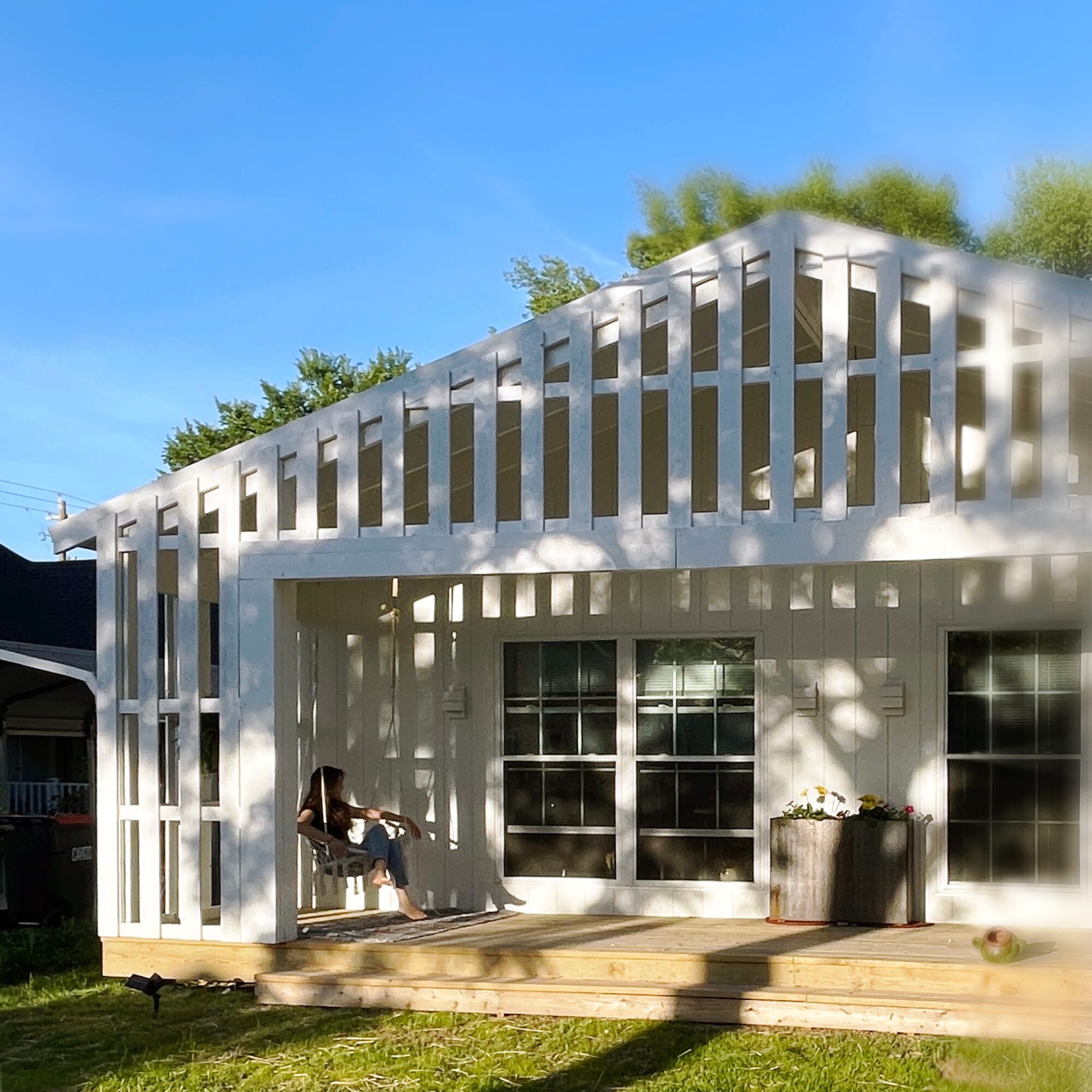 White house with large windows and a wooden deck, person sitting on a bench outside, blue sky and trees in background.