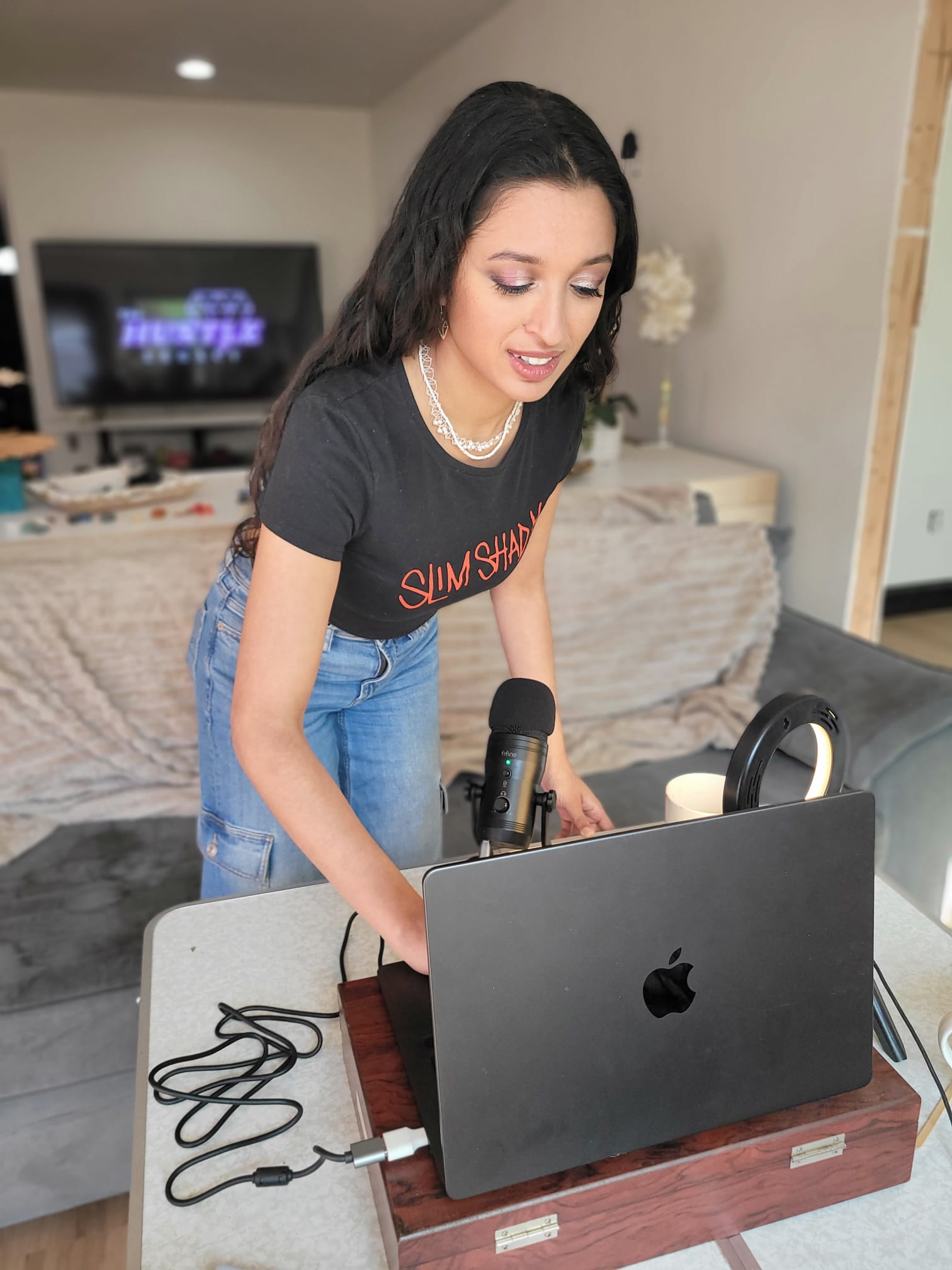 Woman with long dark hair wearing a black t-shirt and jeans, standing at a table with a laptop and microphone.
