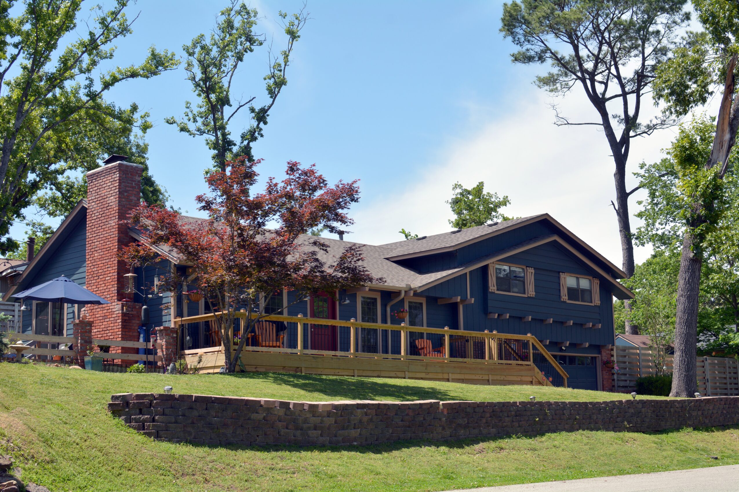 House with a dark exterior, brick chimney, surrounded by trees and a grassy yard, under a blue sky.