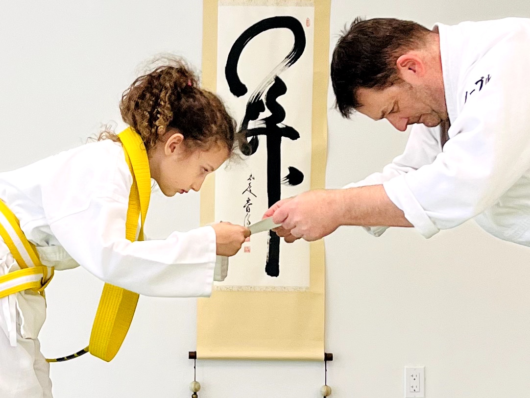 A young girl and an adult man in martial arts uniforms exchange a certificate in front of a calligraphy scroll.