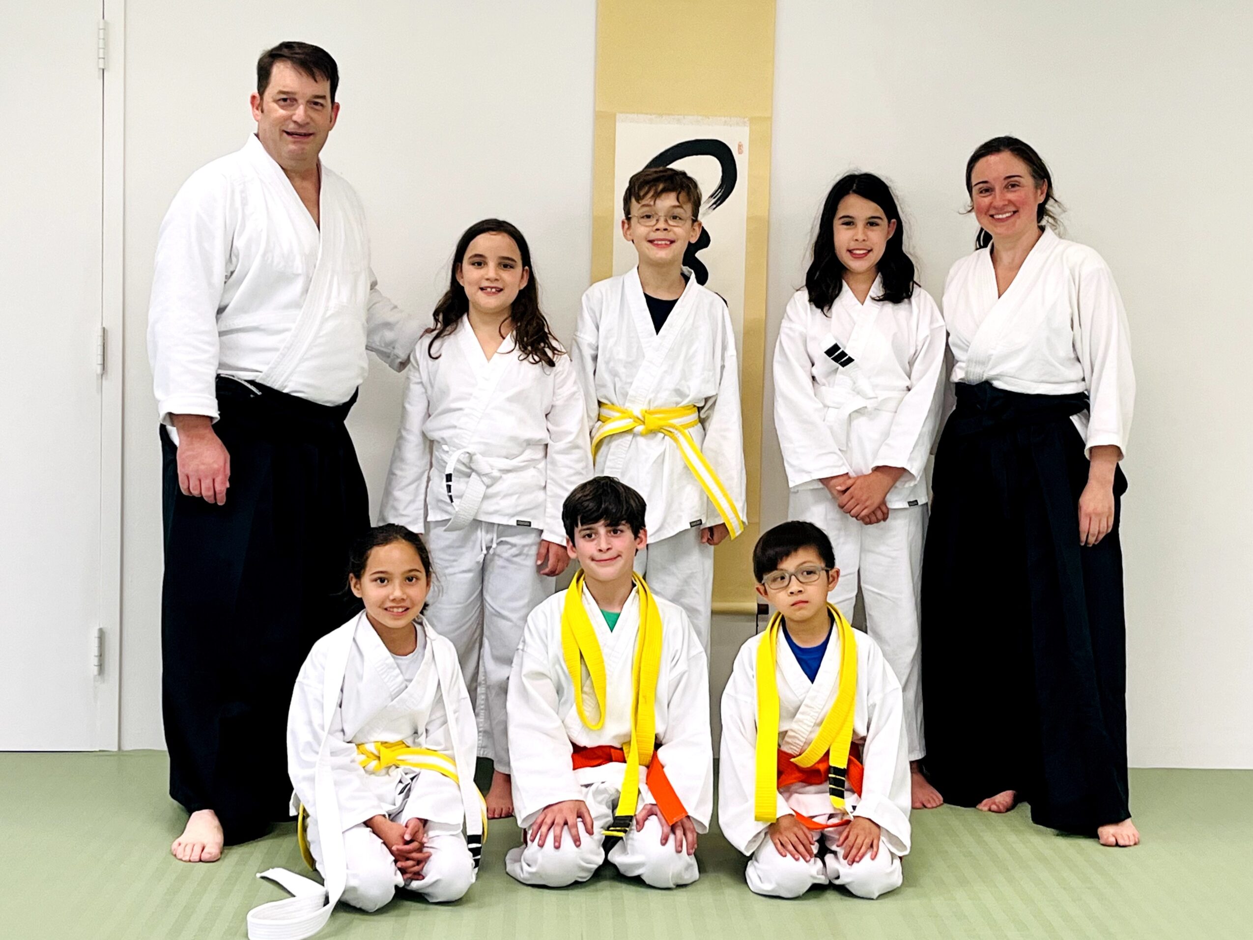 Group of children and two adults in martial arts uniforms standing indoors, smiling, with a yellow belt on one child.
