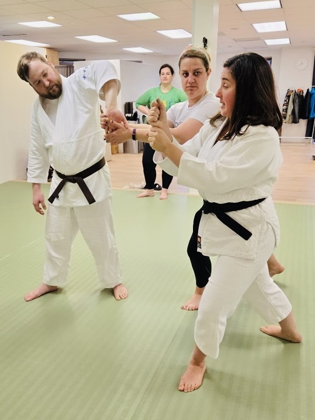 Two women in martial arts uniforms practicing a technique with a male instructor in a dojo, others observing in the background.