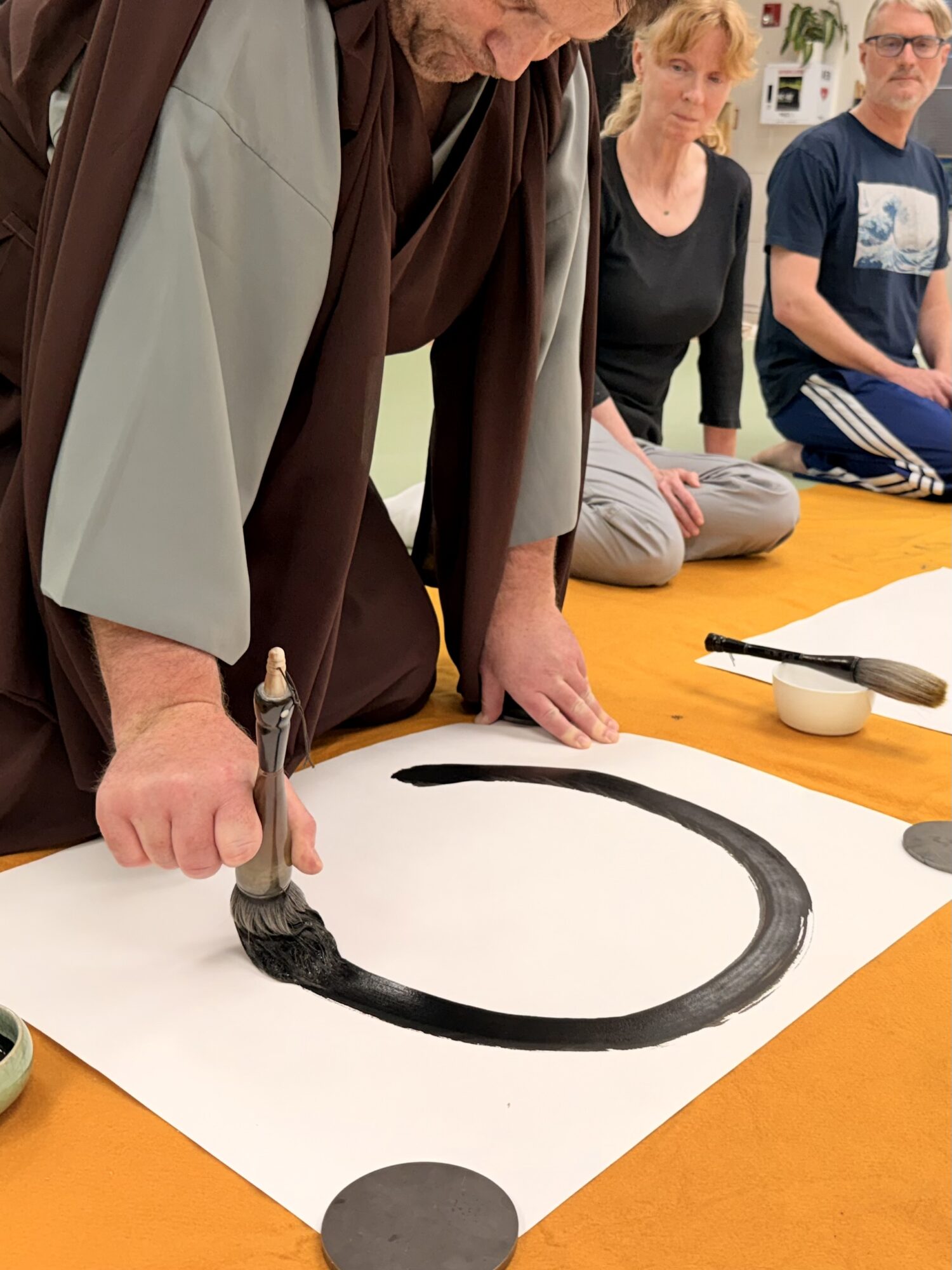 Person painting a large black circle on paper with a brush, others sitting nearby, on an orange table.