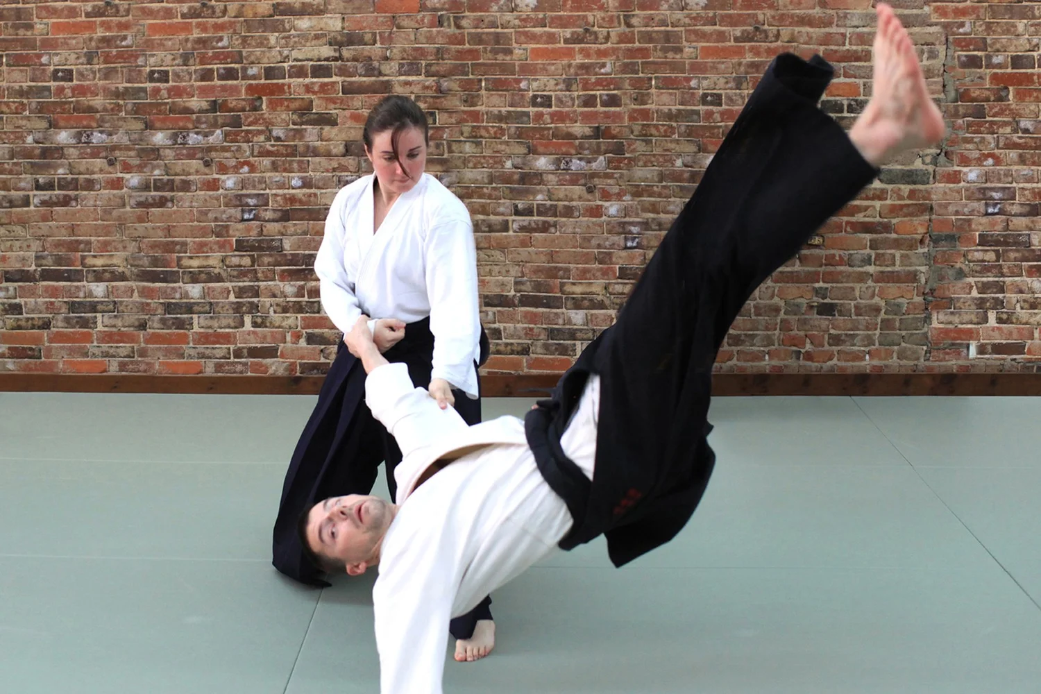 Two people practicing martial arts on a mat against a brick wall, one person in a white gi performing a move.