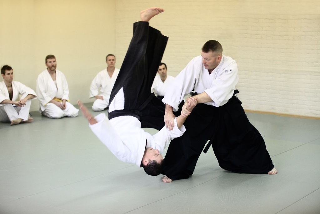A martial arts demonstration with two practitioners, one executing a throw, others watching in a dojo.