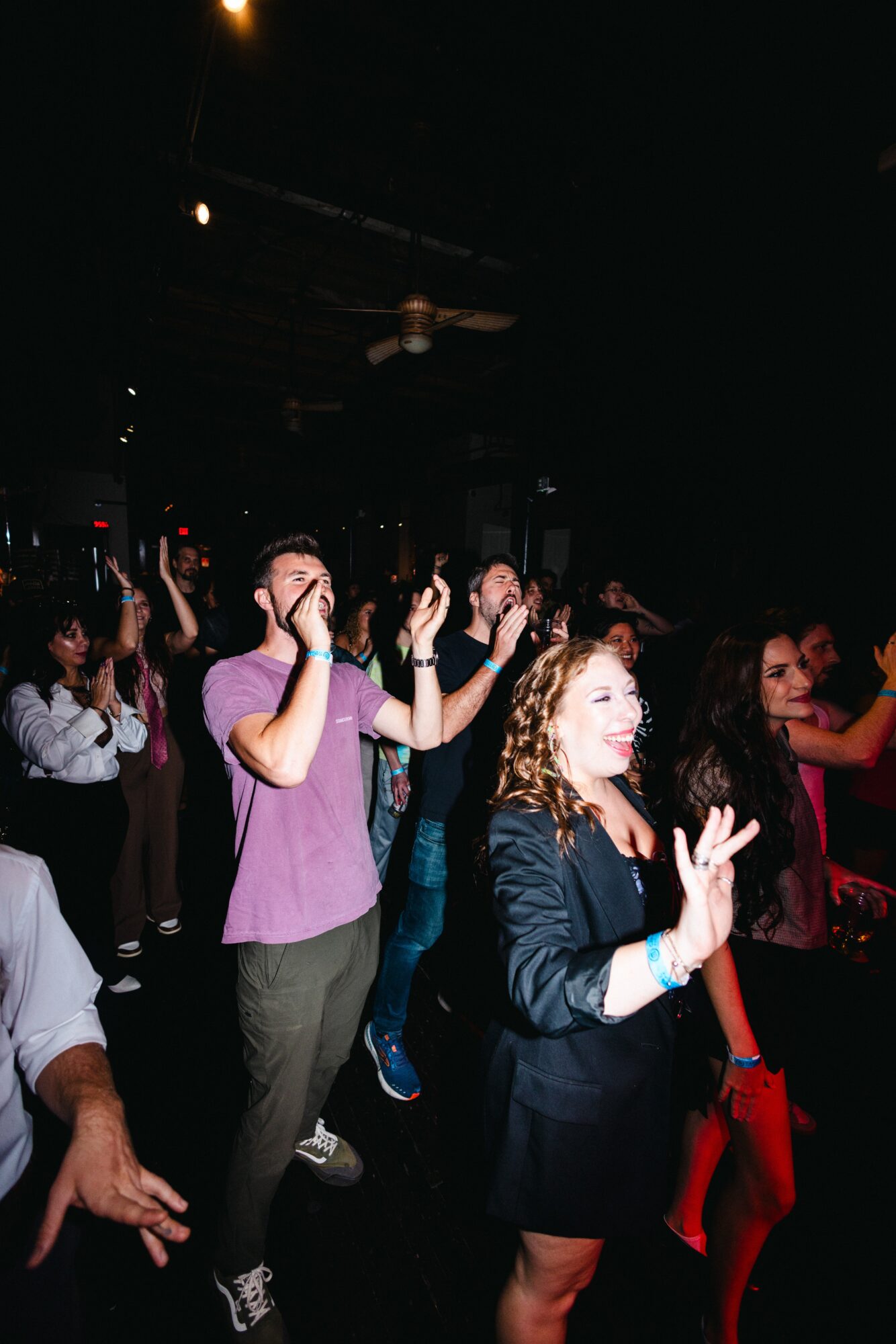 People dancing and enjoying music in a dark indoor setting with some raised hands and smiling faces.