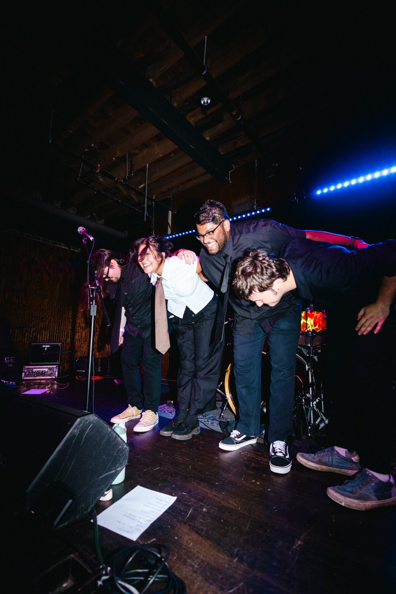 Group of five people on stage bowing after a performance, with musical instruments and stage lighting visible.