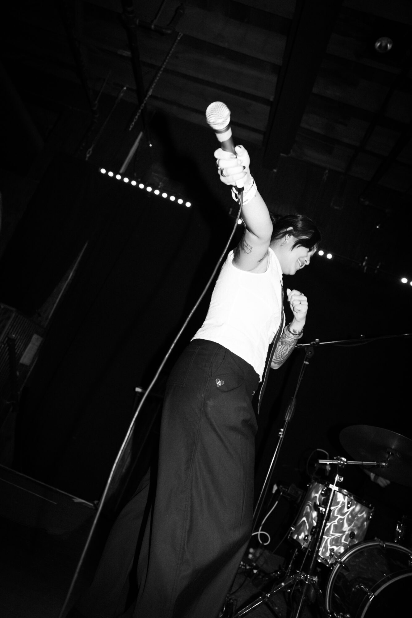 Person holding microphone on stage, wearing a white top and dark pants, with a drum set in the background.