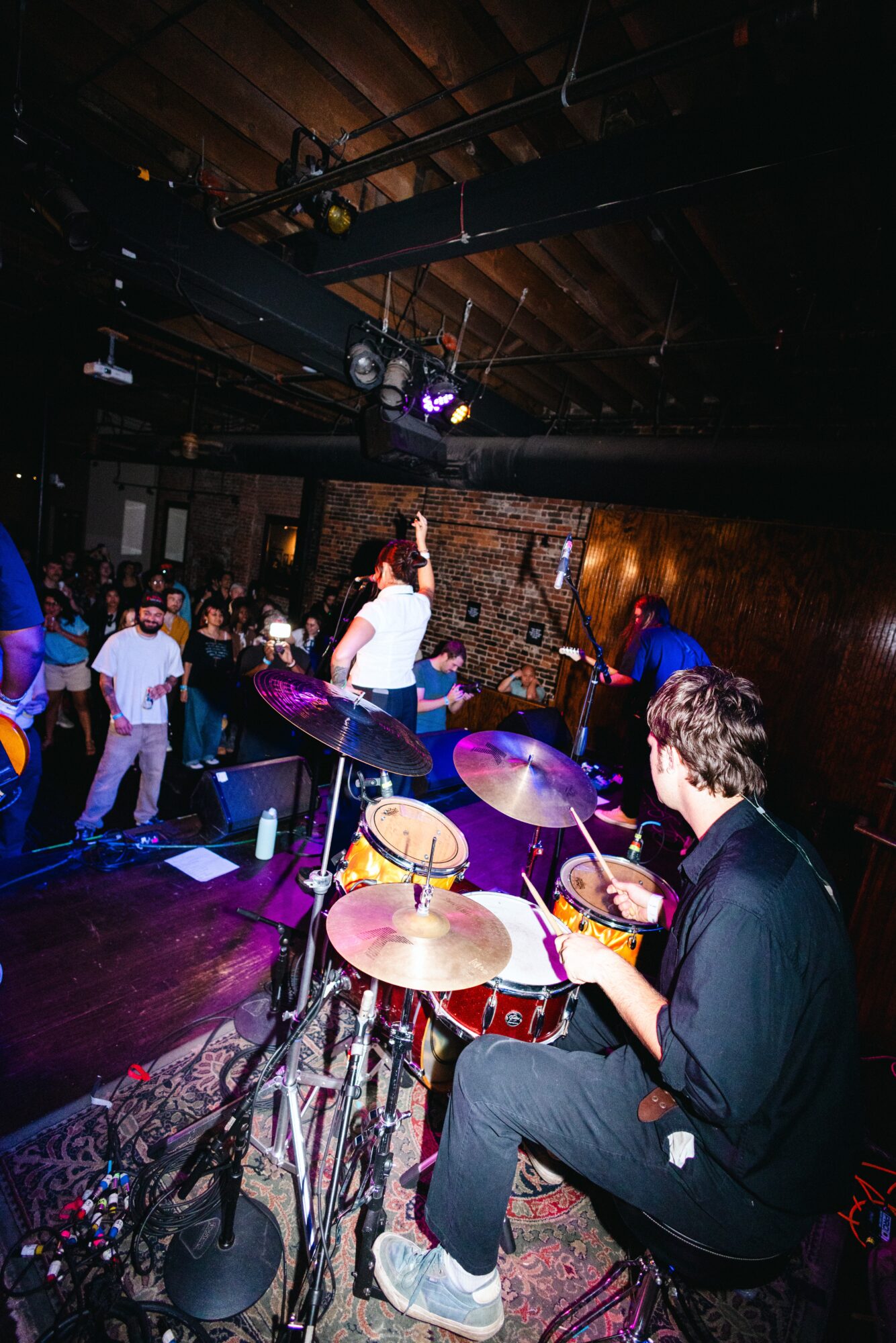 Musician playing drums on stage with people dancing in a dimly lit venue, wooden ceiling, and colorful lighting.