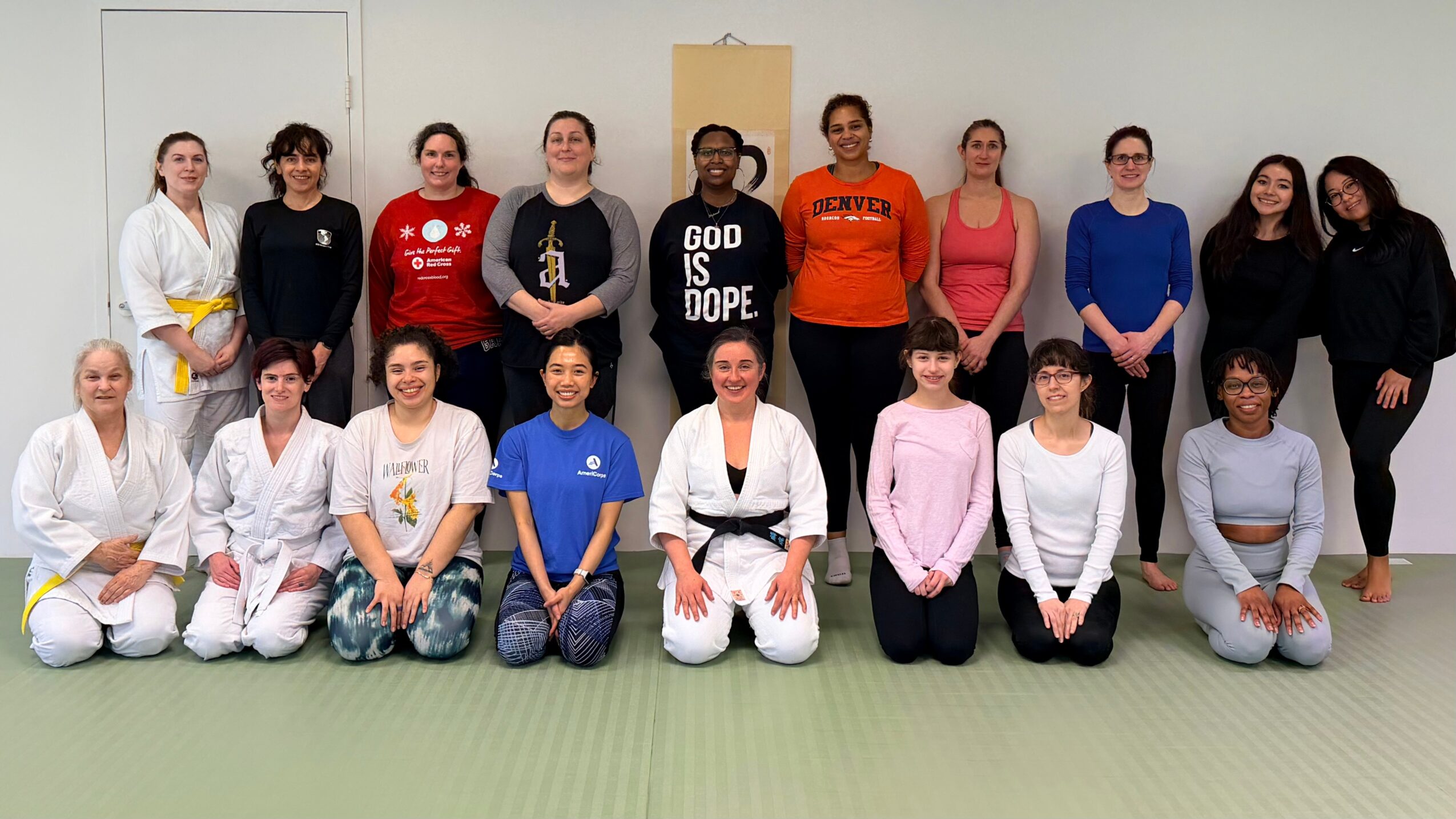 Group of 16 people, some in martial arts uniforms, posing indoors on a green mat, with a yellow banner in the background.