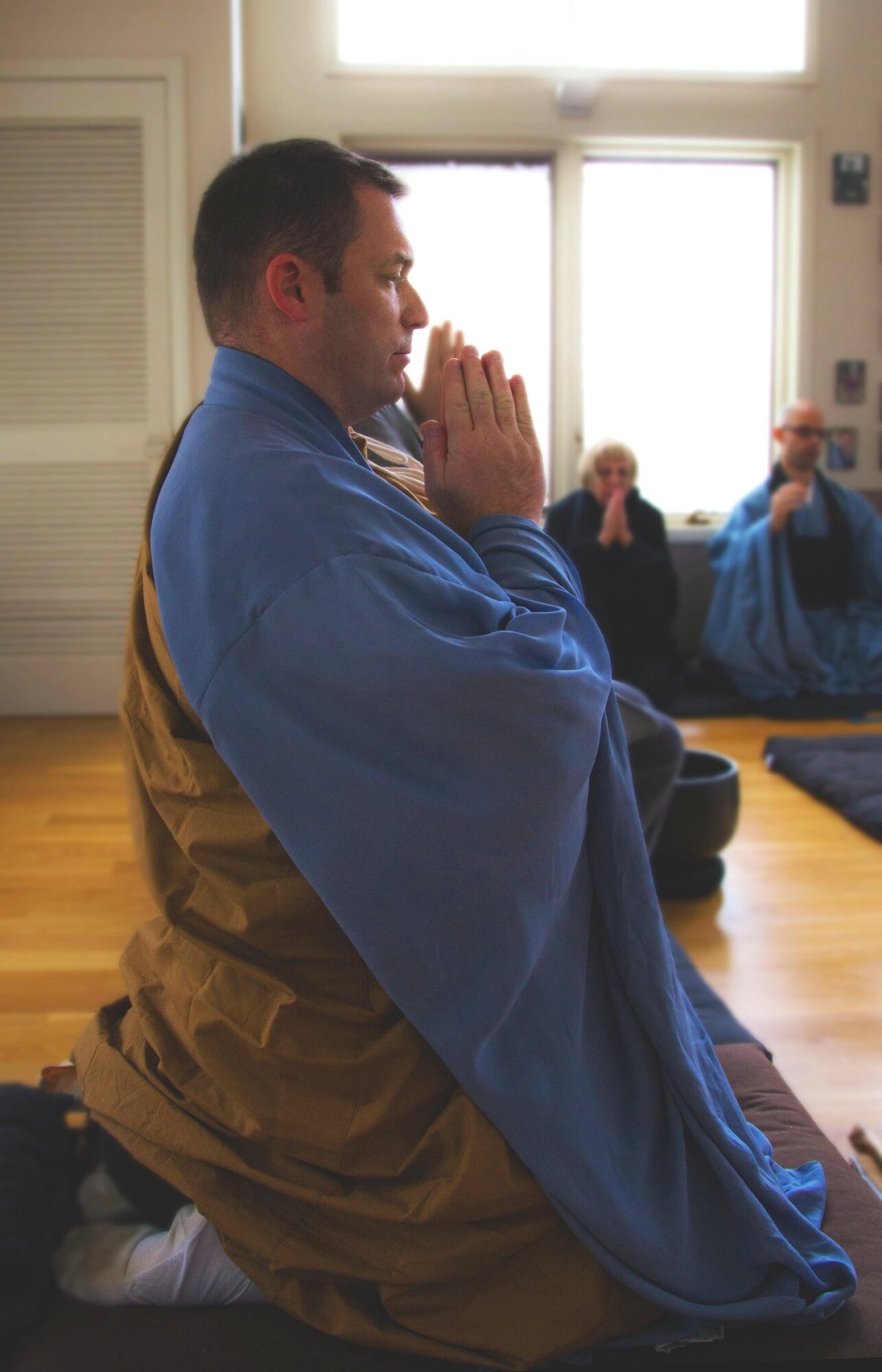Person kneeling with hands in prayer, wearing a blue and brown robe, in a room with others sitting on the floor.