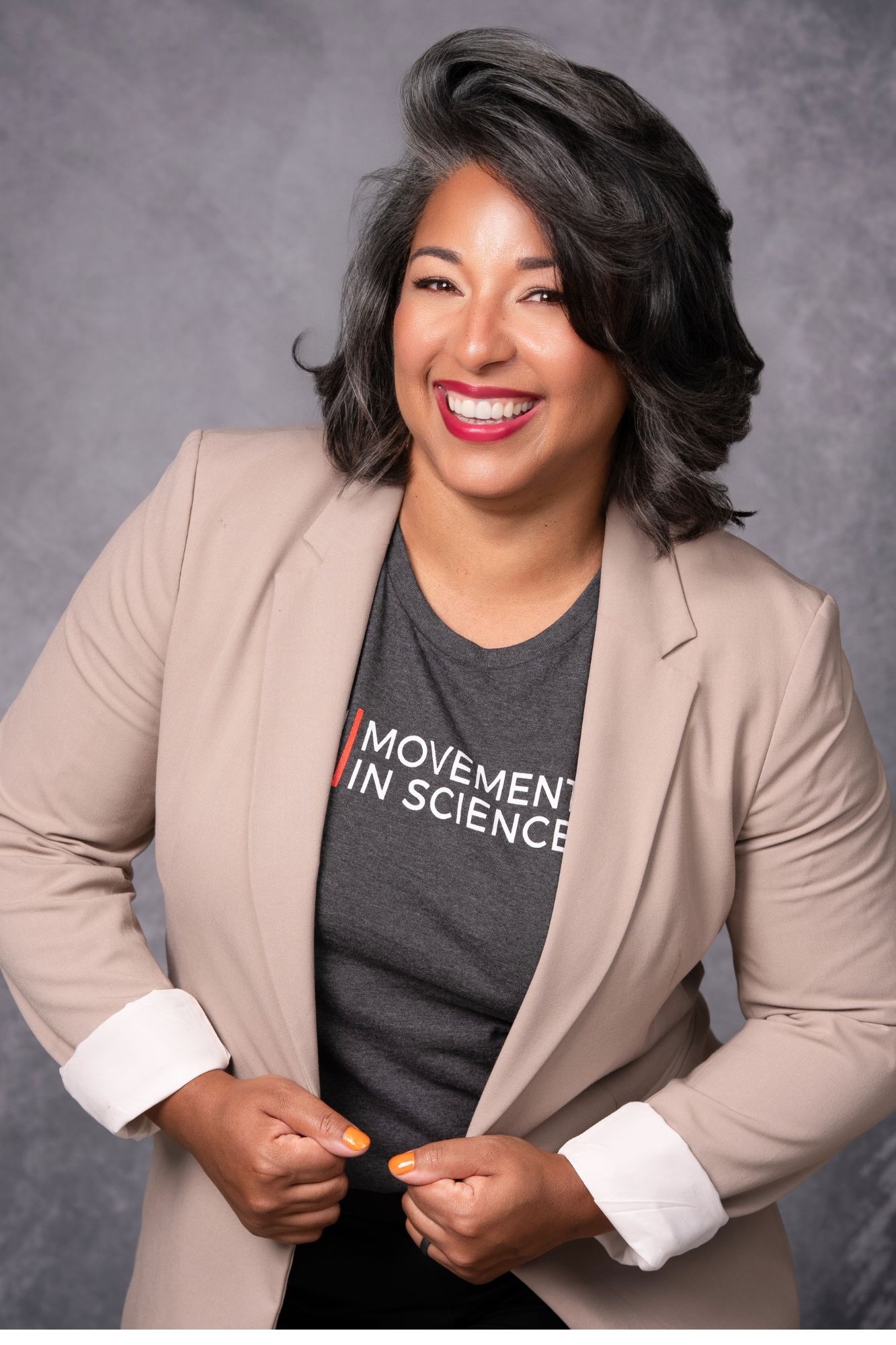 Smiling woman with shoulder-length dark hair, wearing a beige blazer over a gray T-shirt, against a gray background.