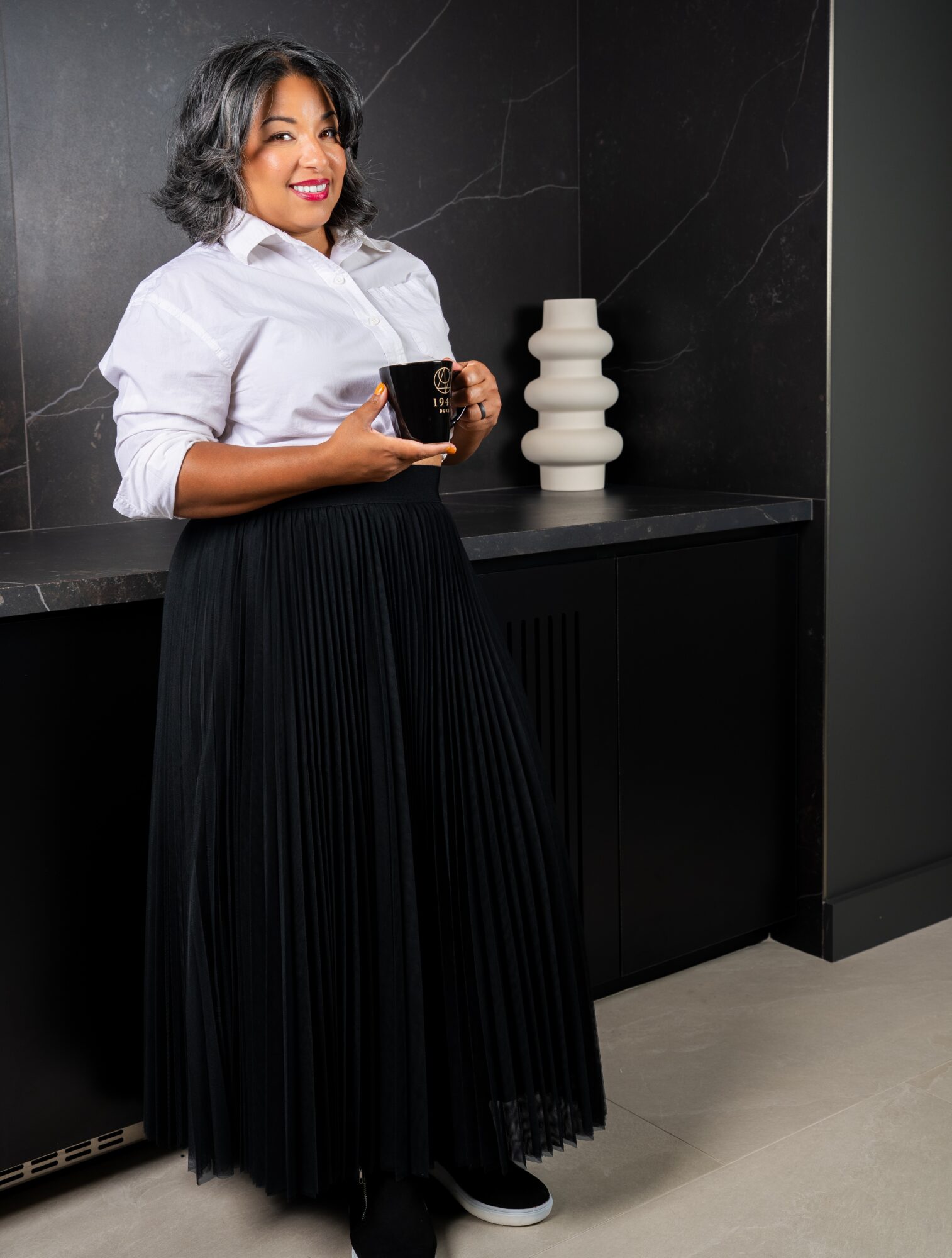 Woman with short curly hair holding a cup, standing in front of a black wall with a decorative white vase, smiling.