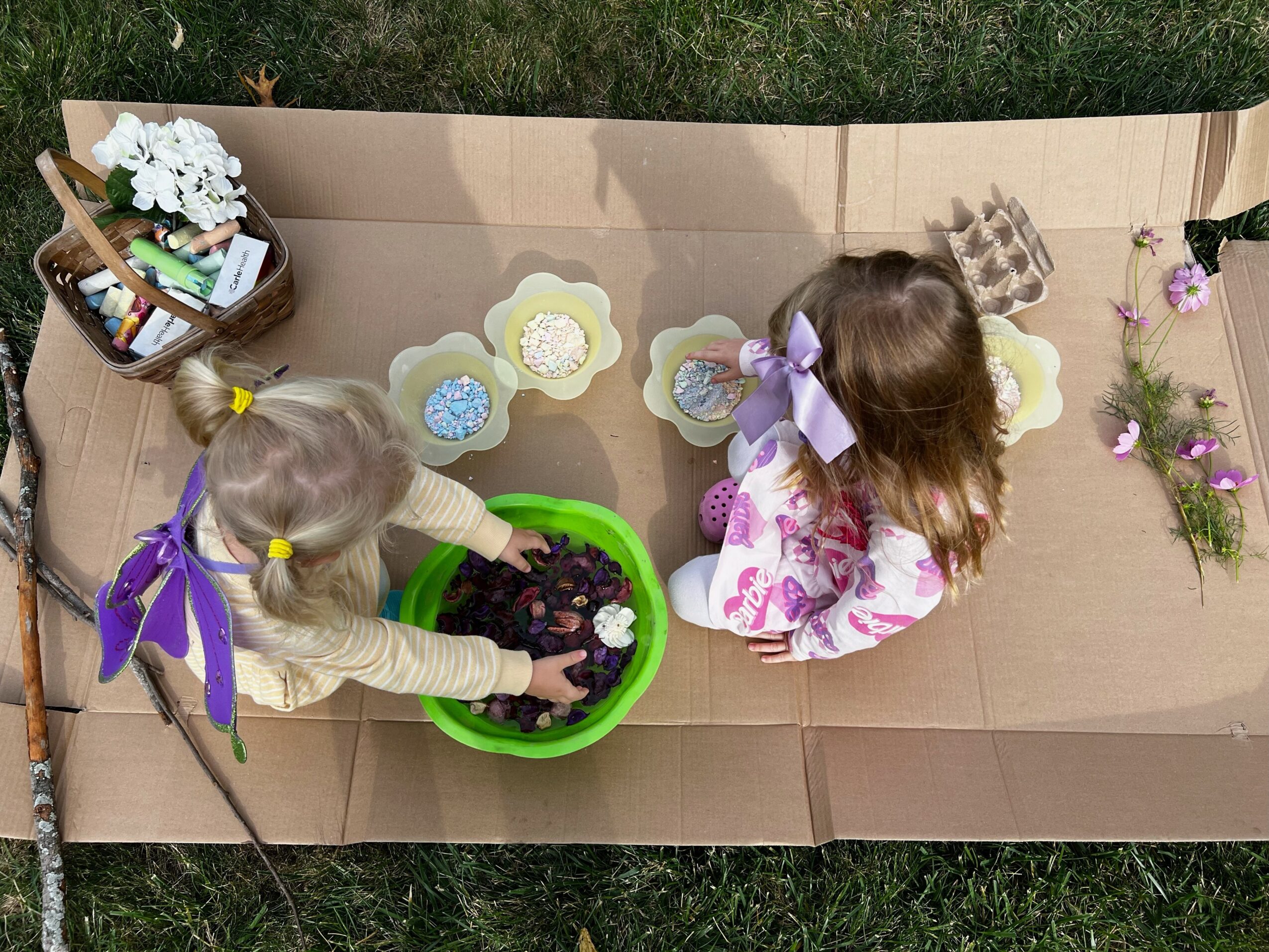 Two children planting flowers in a green container, surrounded by gardening supplies and flower decorations on a cardboard surface.
