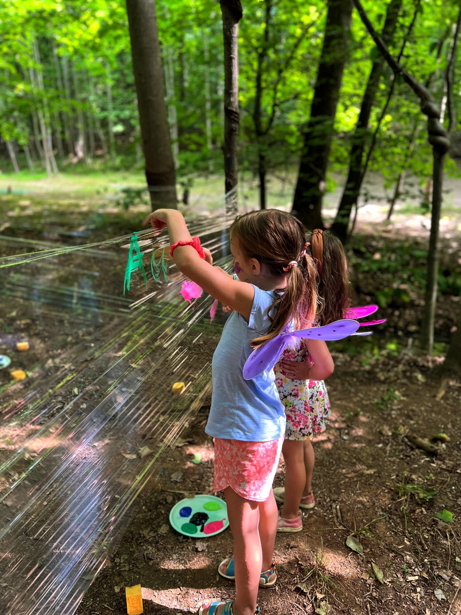 Two children playing with a water spray in a forest, one girl in a blue shirt and pink shorts, the other in a floral dress, holding a purple toy, with trees in the background.