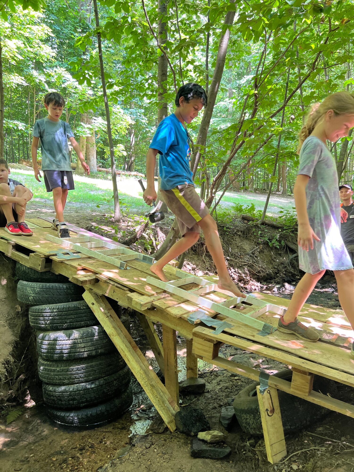Children walking on a wooden bridge in a forested area with green trees and sunlight filtering through leaves.