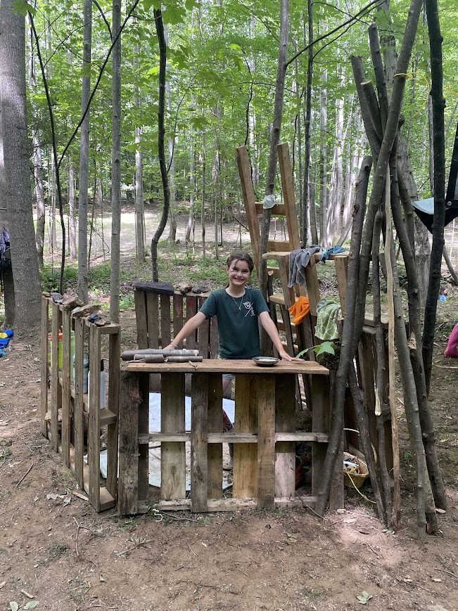 Child standing inside a wooden play area surrounded by trees in a forest, smiling, with a ladder and wooden structures nearby.