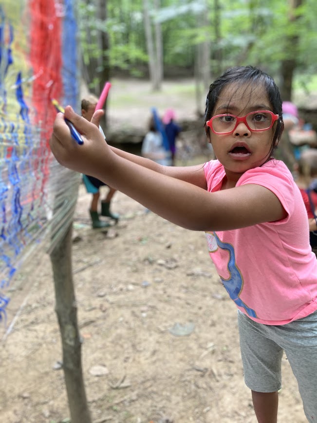 Young girl with glasses and pink shirt drawing on a colorful outdoor wall, in a wooded area.