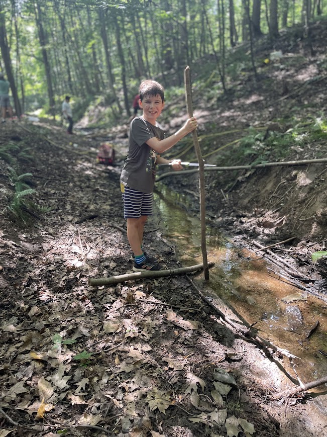 Child standing on a makeshift wooden bridge over a small creek in a forest, holding a stick, surrounded by trees and leaves.
