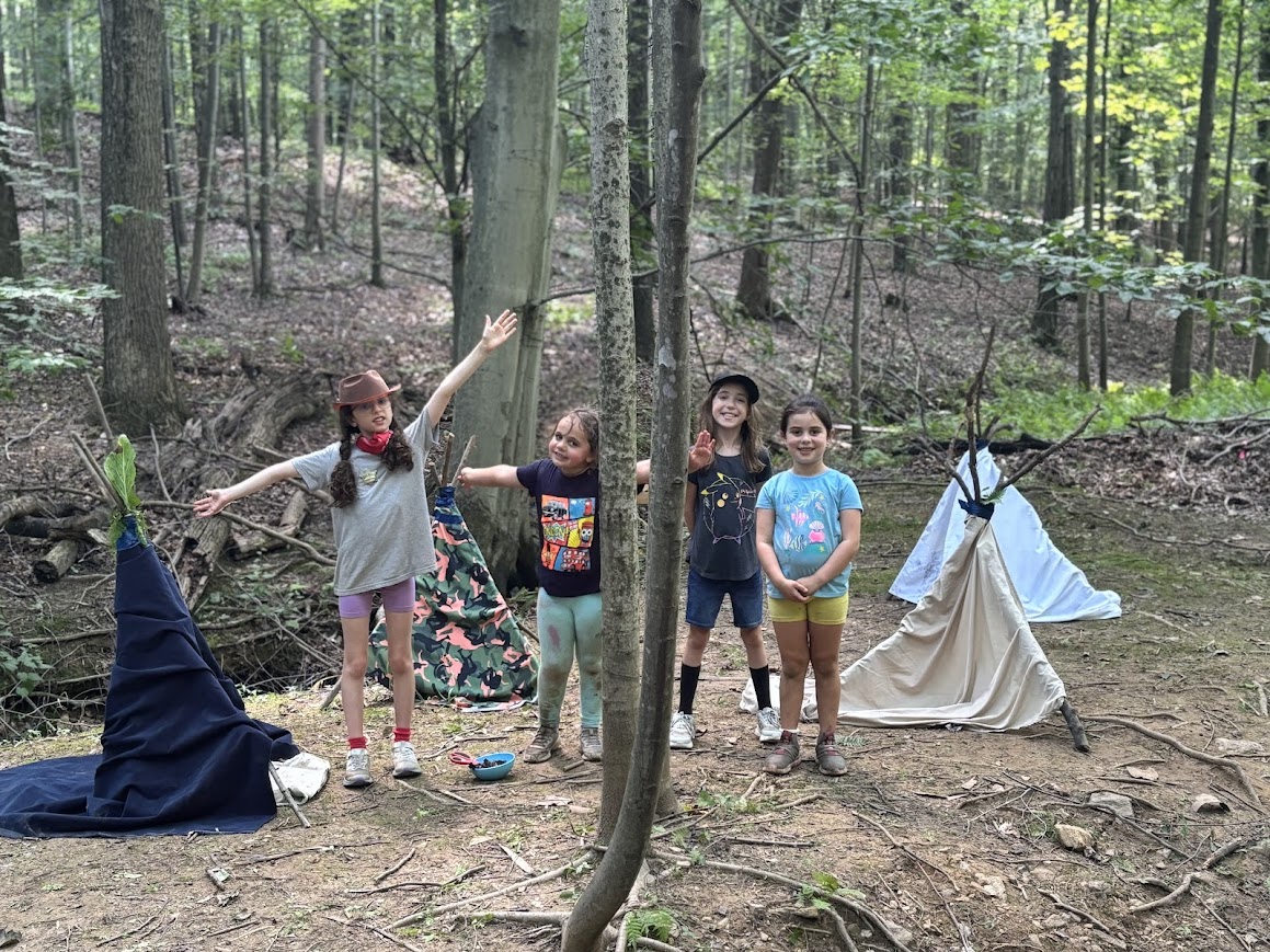 Five children stand in a forest campsite with tents, trees, and camping gear, smiling and posing for the photo.