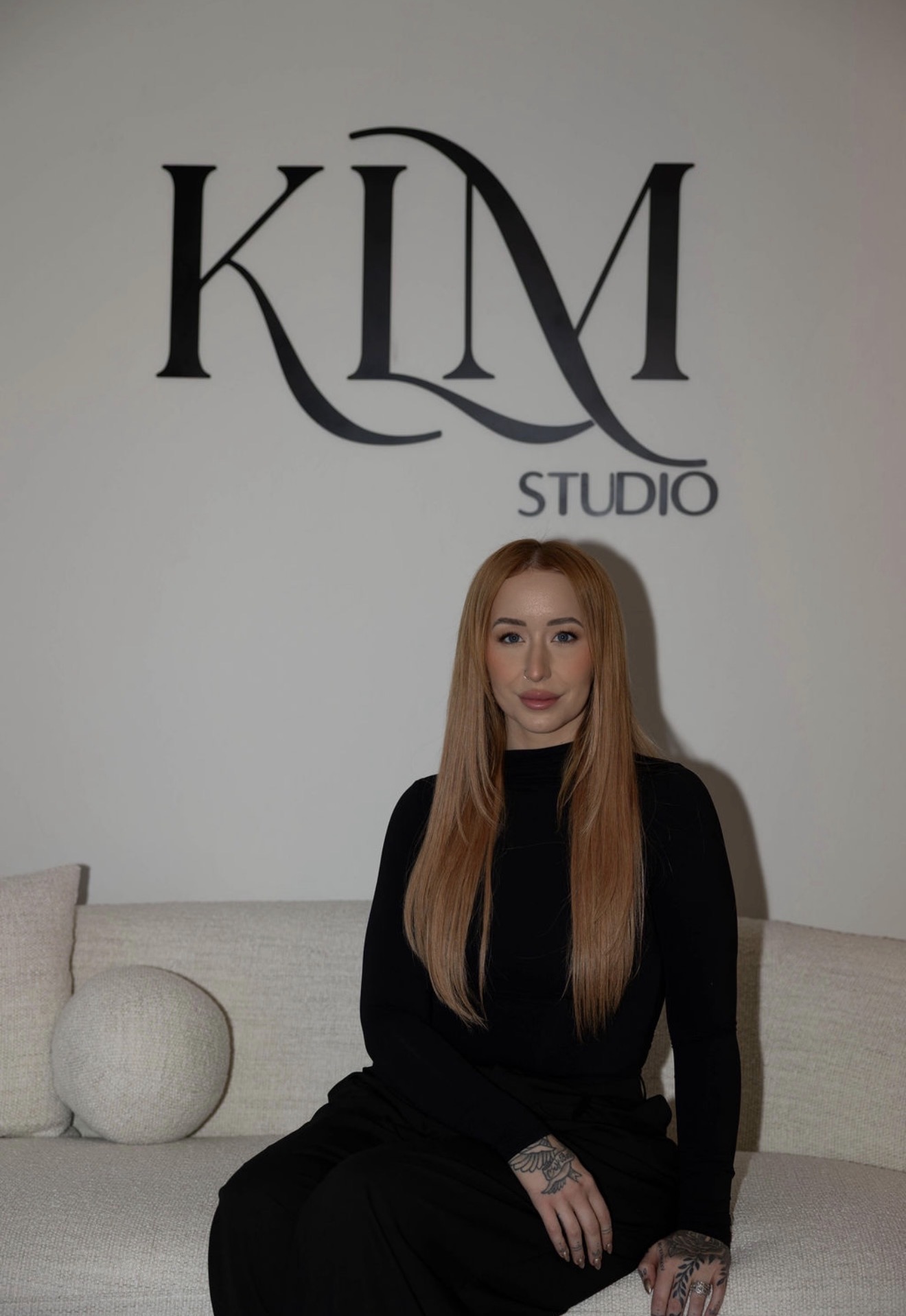 A woman with long hair sitting on a sofa in front of a wall with 'KLM Studio' logo.