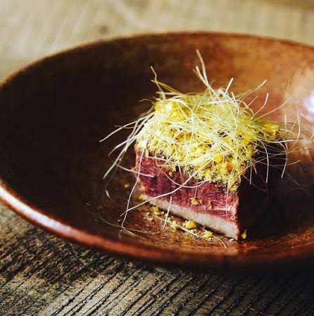 A piece of raw meat with yellowish fibrous material on top, in a brown bowl on a wooden surface.