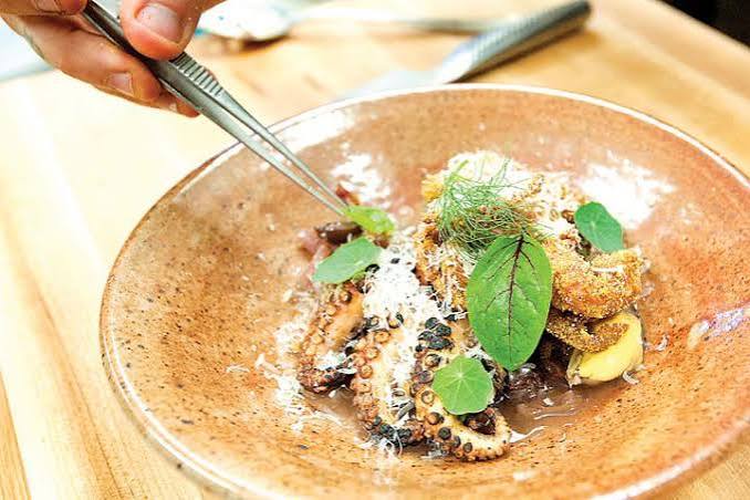 Person garnishing a dish with herbs and grated cheese on a brown plate, with a hand holding tweezers.