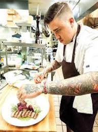 Person with tattoos preparing food in a kitchen, focusing on plating meat dishes, wearing a white shirt and apron.