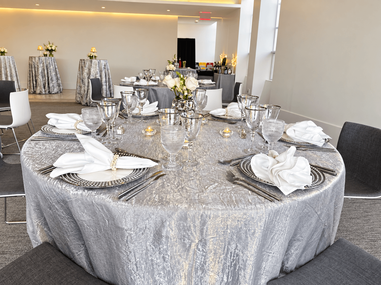 Round table with a silver tablecloth, set with white napkins, glassware, and plates, in a decorated event space.