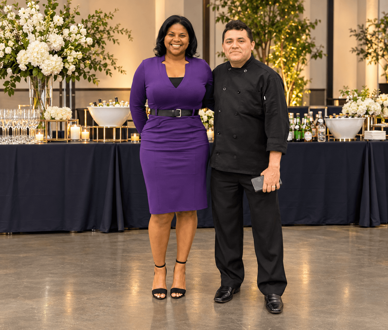 A woman in a purple dress and a man in black attire stand together in front of a decorated table with flowers and drinks.