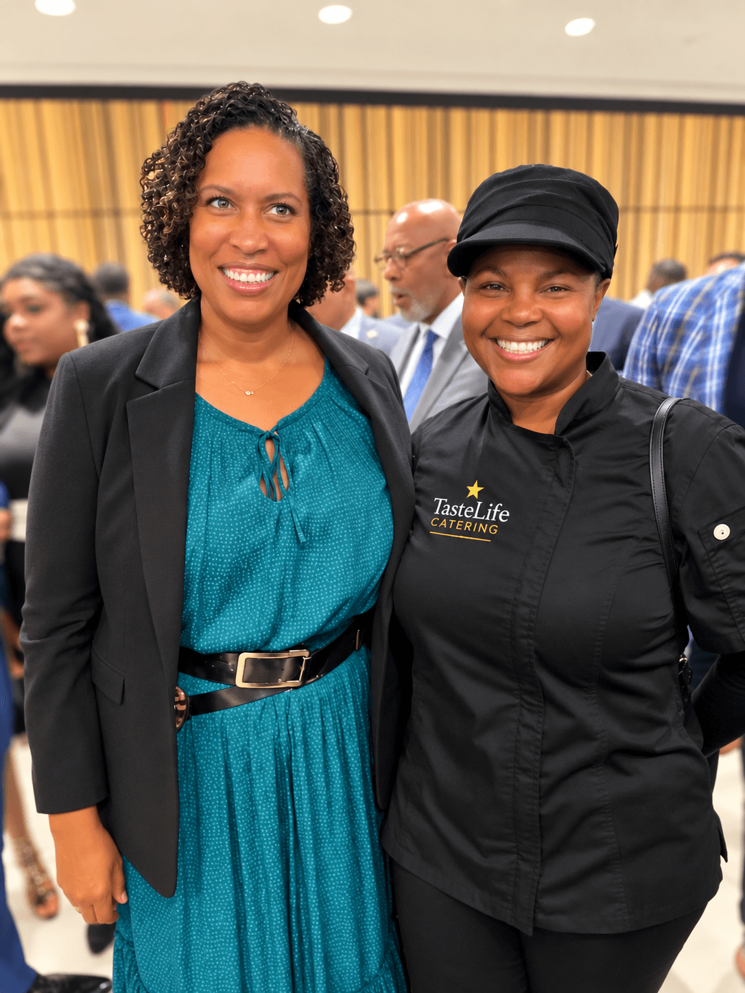 Two women smiling at an indoor event, one in a teal dress and black blazer, the other in a black uniform with a cap.