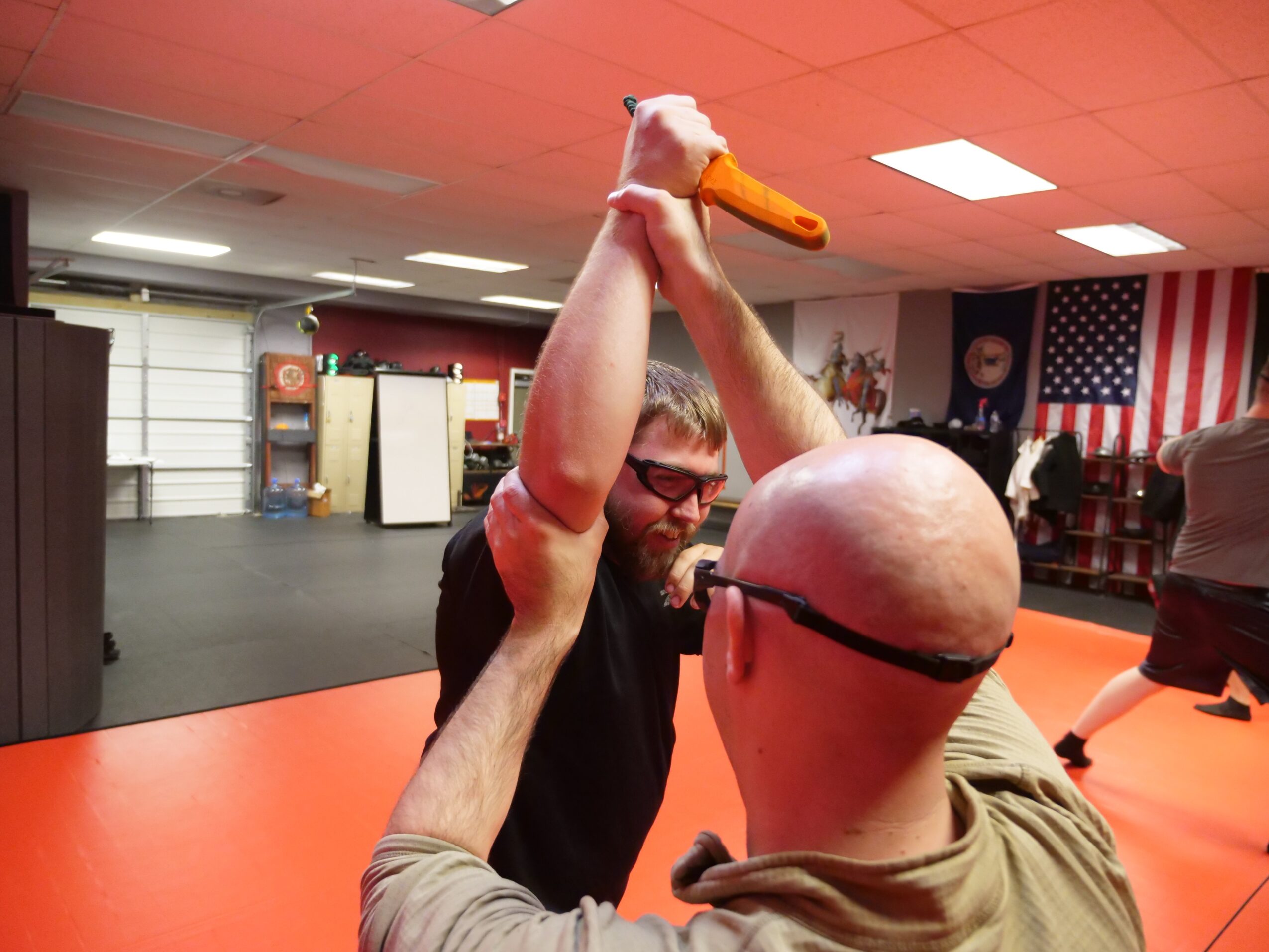 Two men in a martial arts gym, one holding a duck mask, with American flags on the wall behind them.
