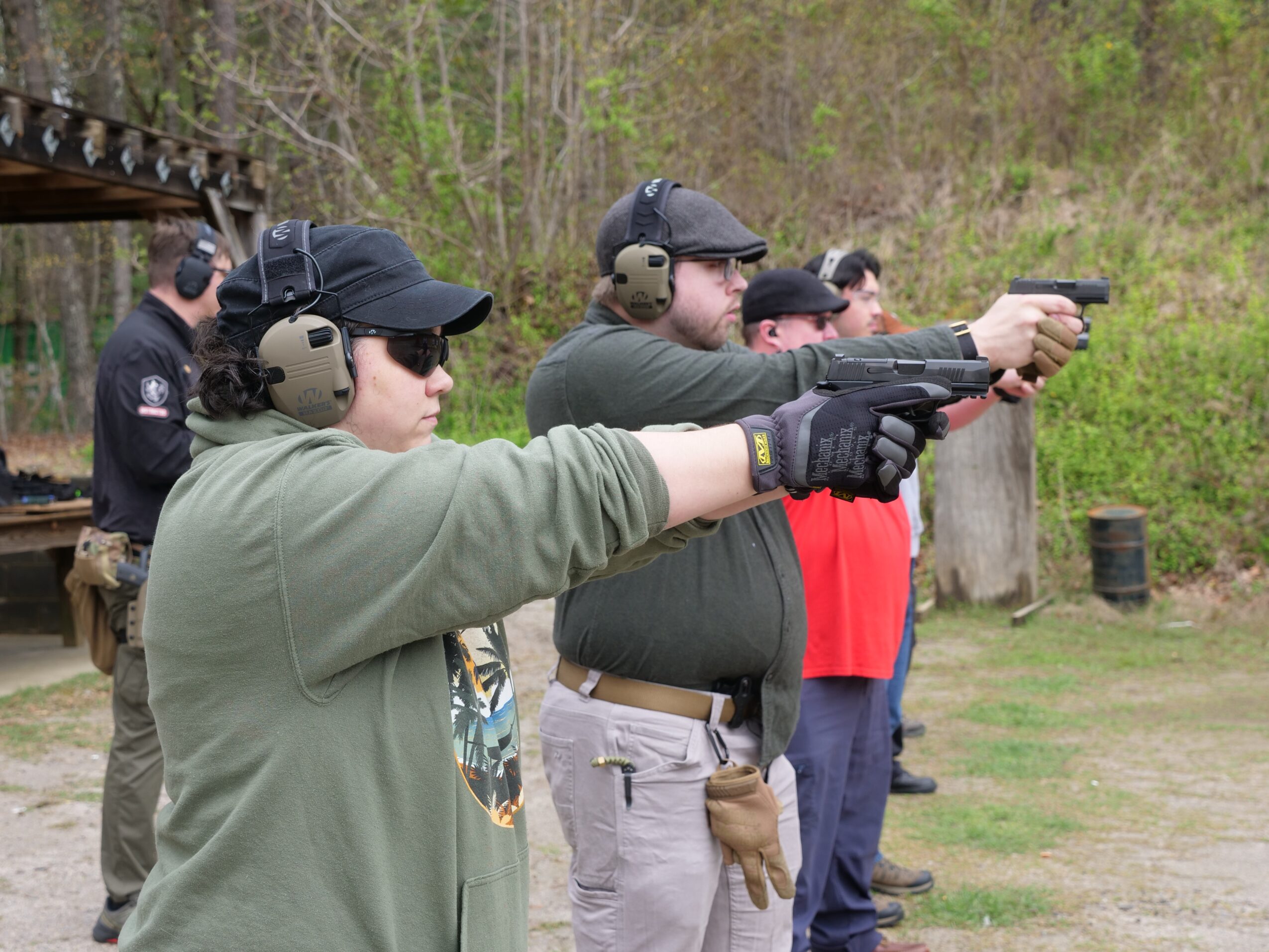 Group of people at outdoor shooting range aiming pistols, wearing ear protection and gloves, with trees and grass in background.
