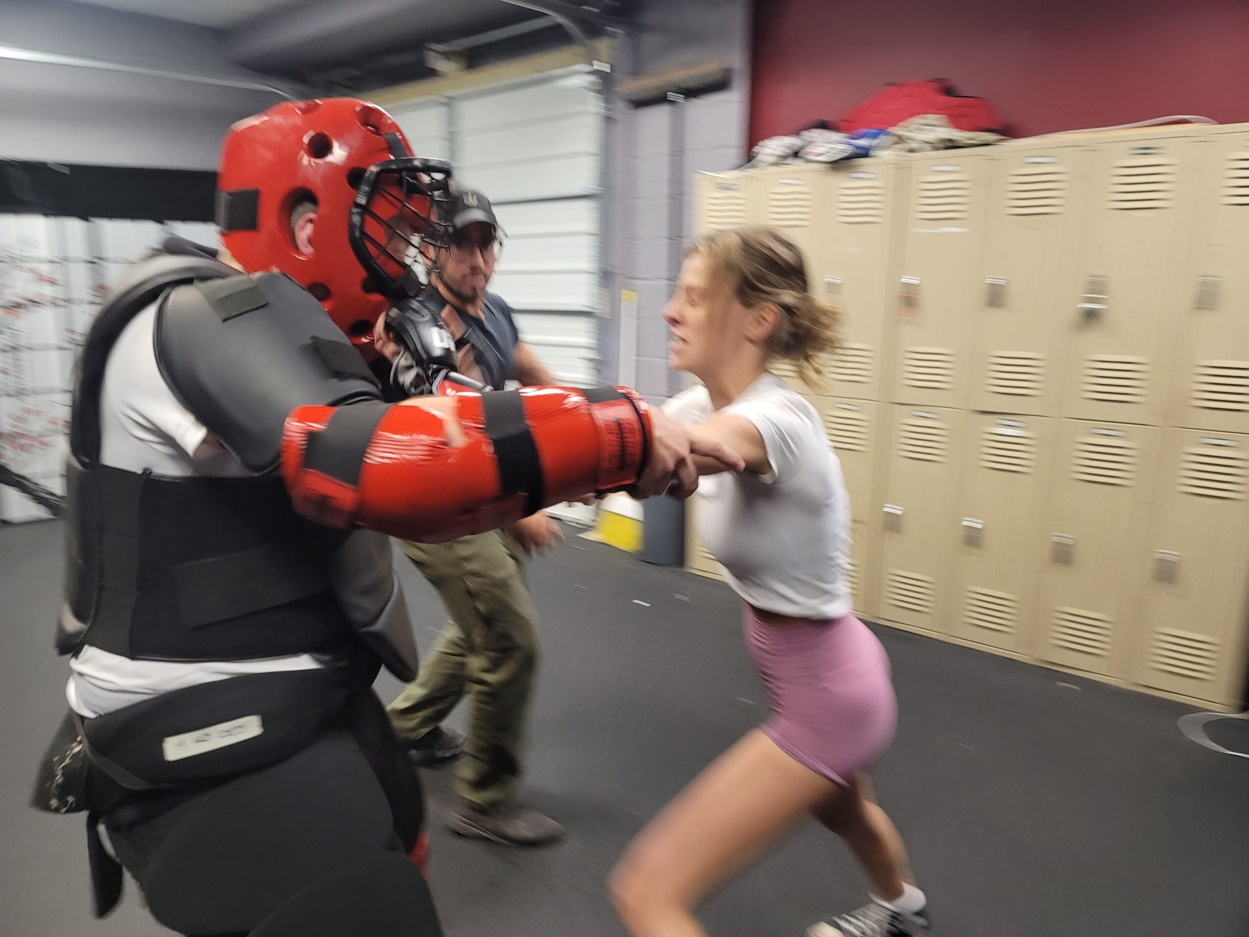 Person lying on the floor with a red helmet, backpack, and gray clothing, next to lockers and a gray wall.