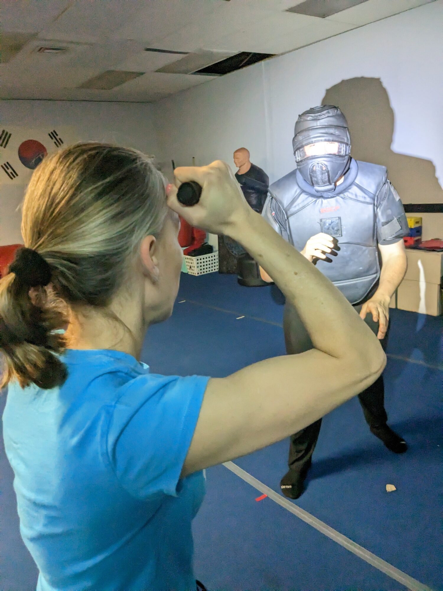 Woman with a ponytail in a blue shirt holds a gun-like object aiming at a person in a virtual reality headset, in an indoor setting.