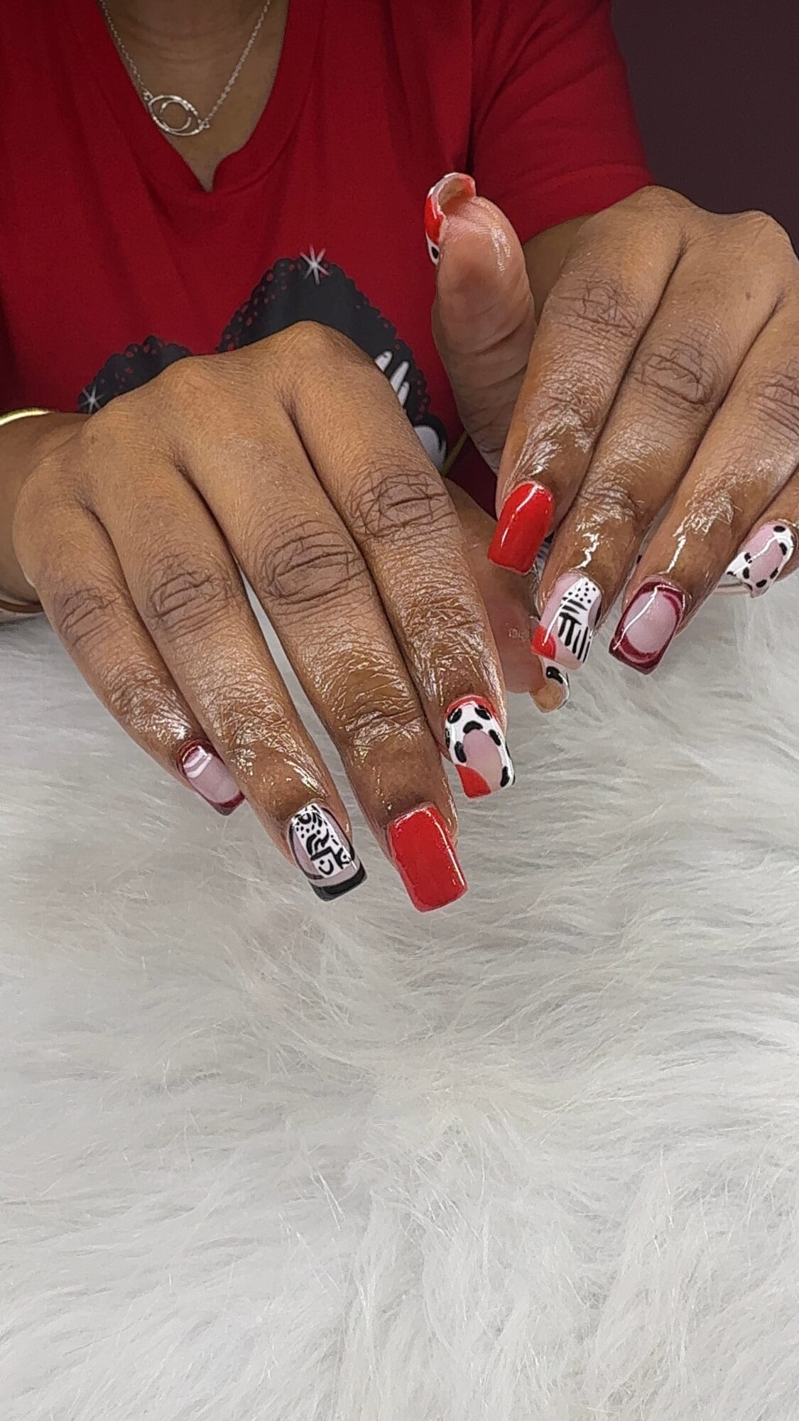 Person's hands with decorated nails resting on a white furry surface, wearing a red top, with a silver necklace visible.
