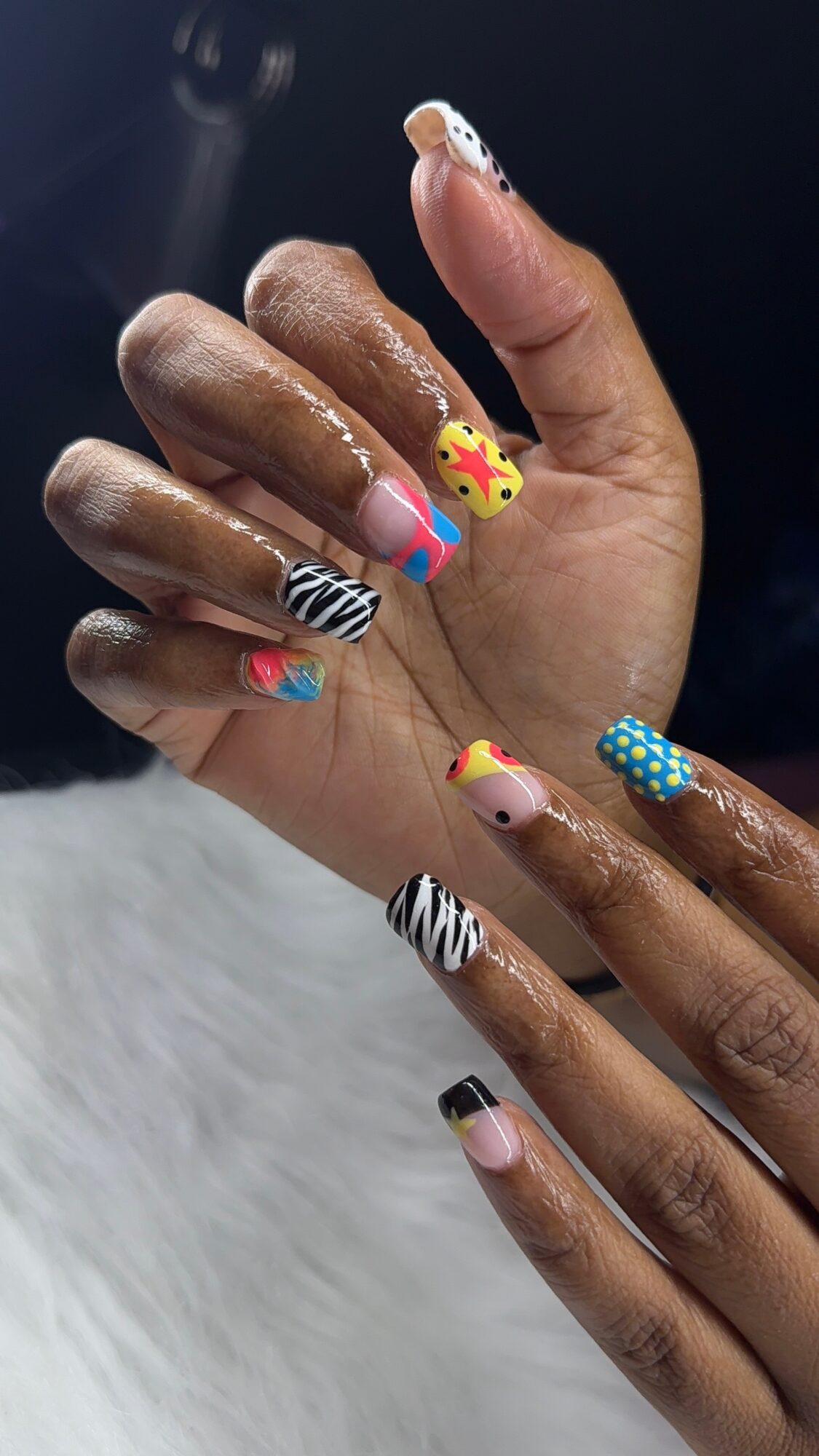 Hand with colorful, decorated nails featuring various patterns and designs, including animals and abstract art, against a dark background.